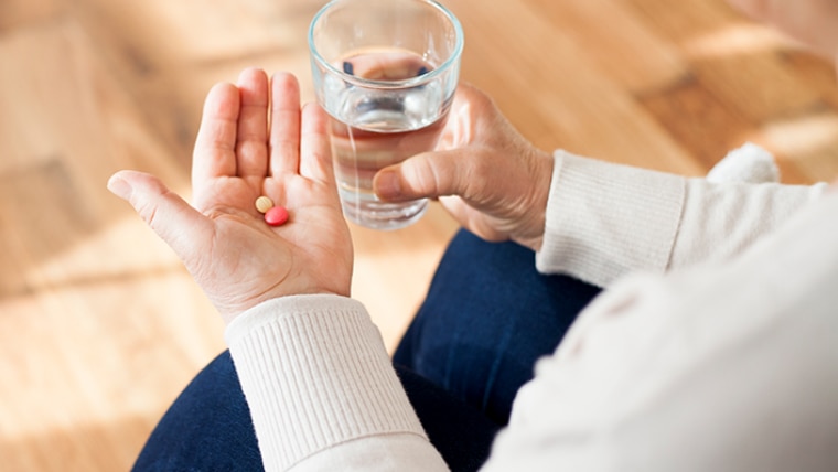 In one hand we see a white pill and an orange pill; in the other hand we see a glass of water.