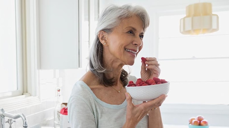A smiling woman stands near her sink with a bowl of raspberries in hand.