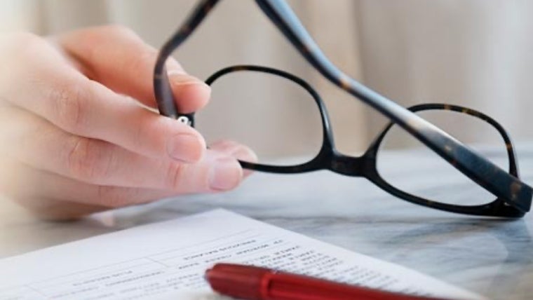 A hand holds the temple of a pair of black glasses over a tabletop filled with papers.