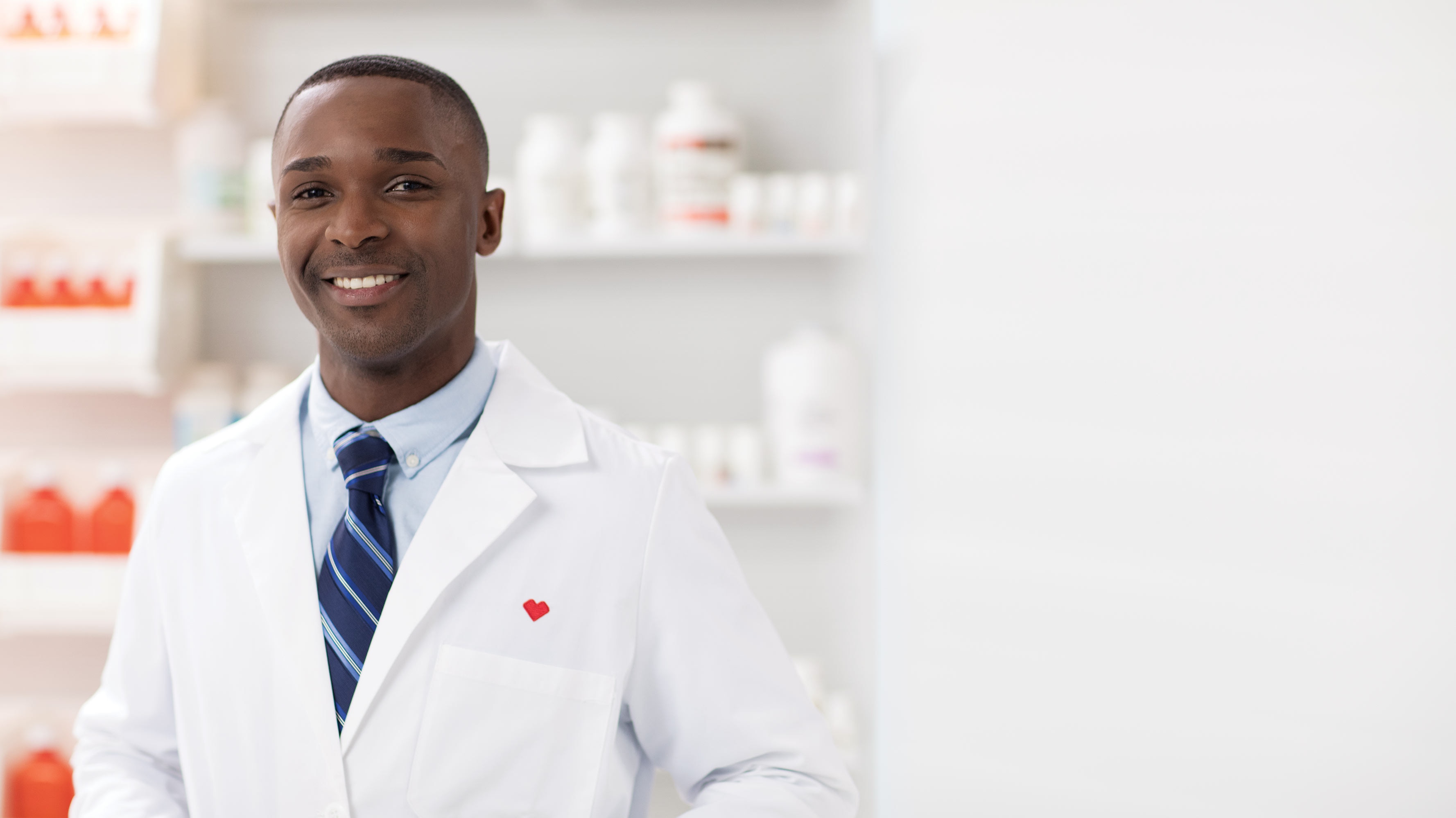 A pharmacist in a while lab coat, embroidered with a red CVS Health heart, smiles while standing behind the counter of a pharmacy.