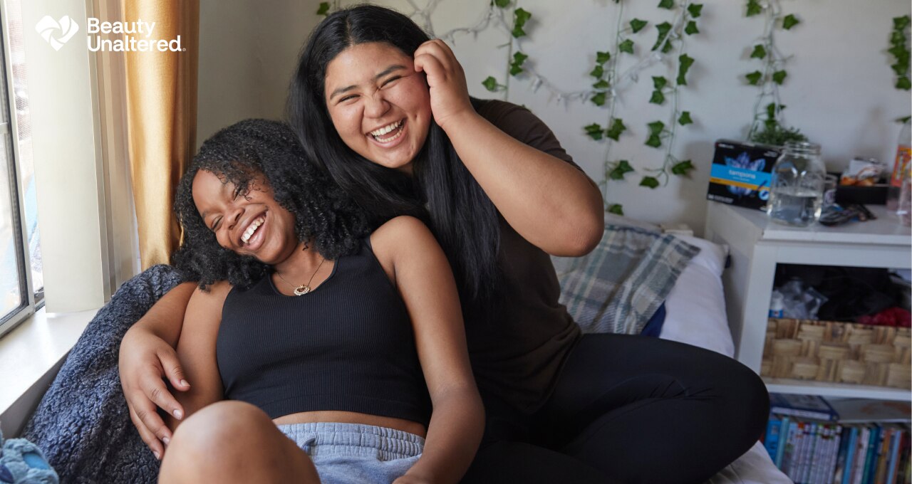 Two young naturally beautiful women of color hang out and laugh together. The logo for beauty unaltered is shown.