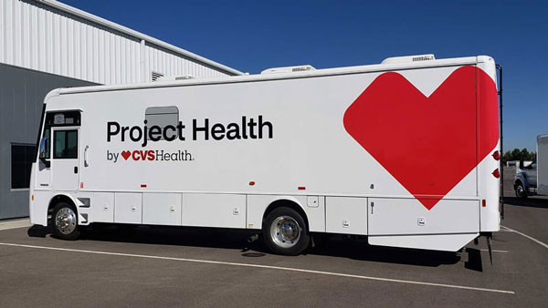 A white Project Health mobile clinic truck by CVS Health parked outside a building, featuring the Project Health logo and a large red heart graphic on the side.