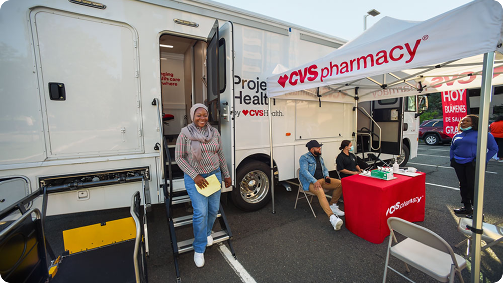 A mobile Project Health clinic truck by CVS Health parked outdoors with its door open, where several people are seated at a CVS Pharmacy booth under a tent, while another person stands on the steps holding papers.