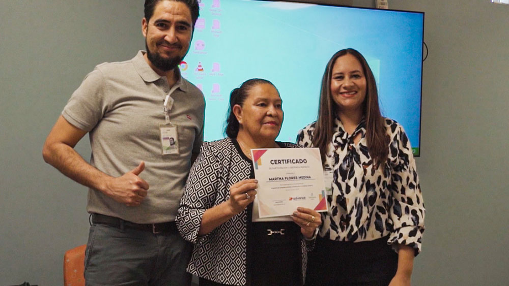 Three people standing indoors in front of a screen, with the person in the center holding a certificate labeled ‘Certificado’ while the two people beside them pose for the photo.