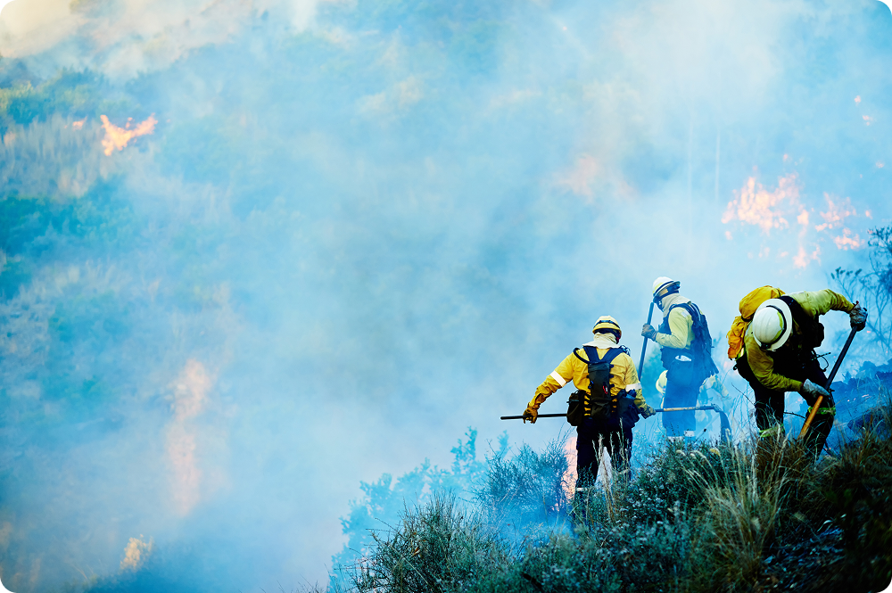 Three firefighters in protective gear moving through smoke and low brush on a hillside during an active wildfire response, with flames visible through the haze in the background.