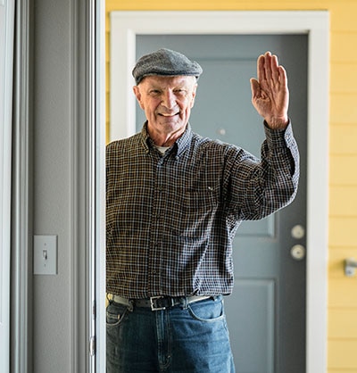 Older man smiling and waving from outside an open door