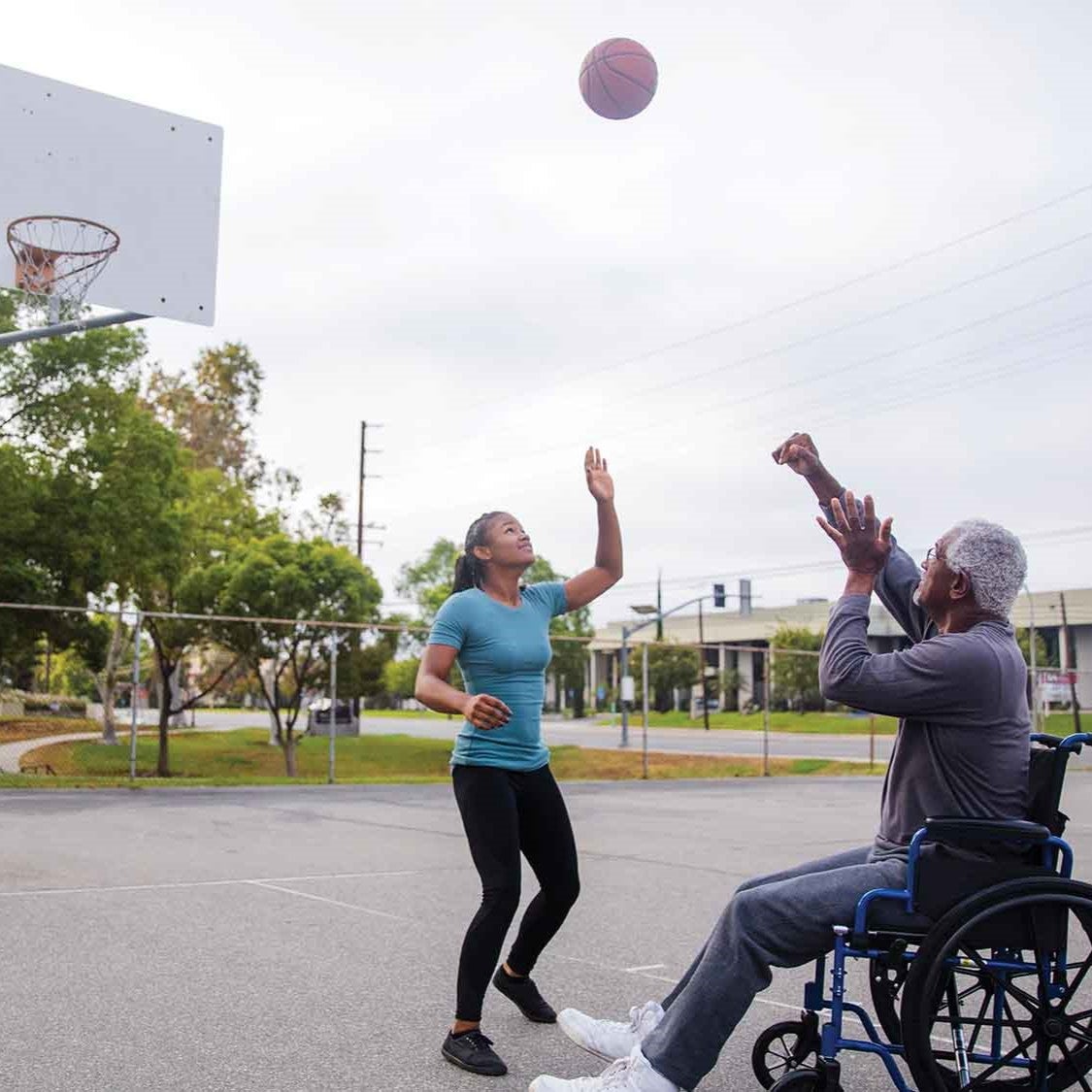 A woman and a man in a wheelchair shoot basketball hoops on a court in a park.