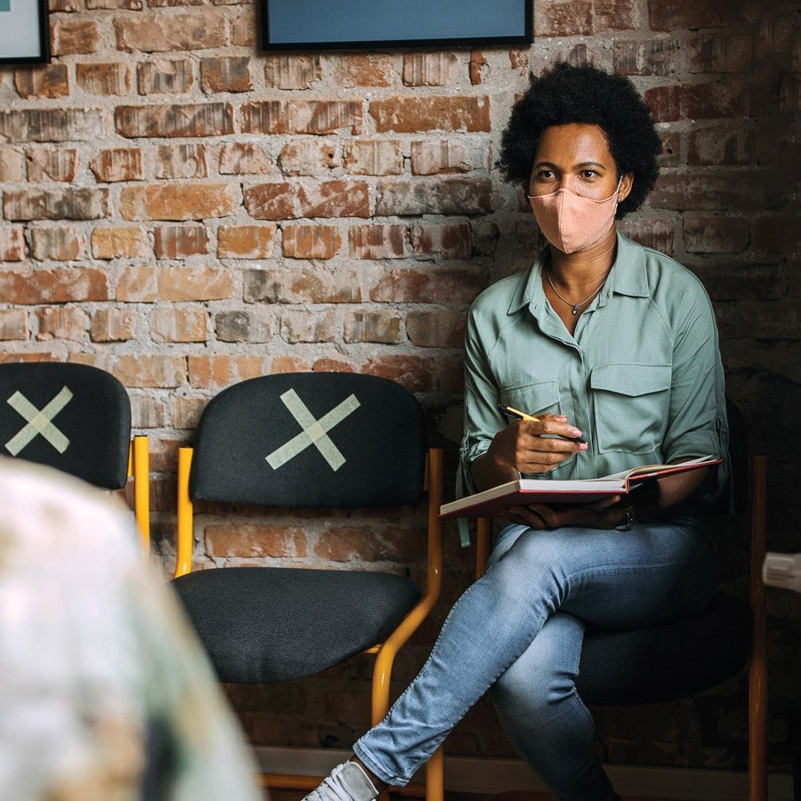 A patient sits in a waiting room filling out a form while talking to a healthcare professional.