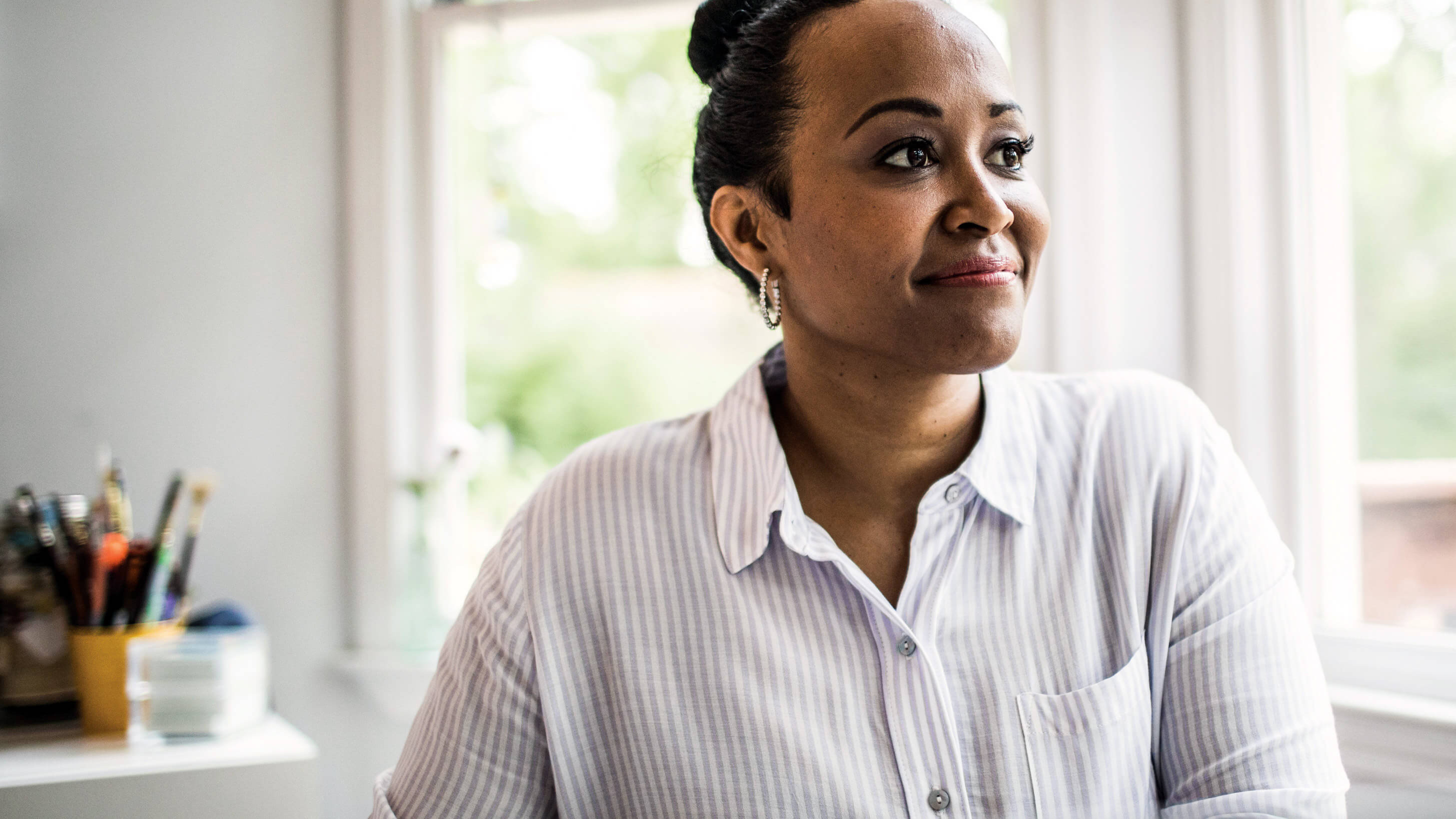 An African-American woman gazes out of a window in her modern home office, smiling.