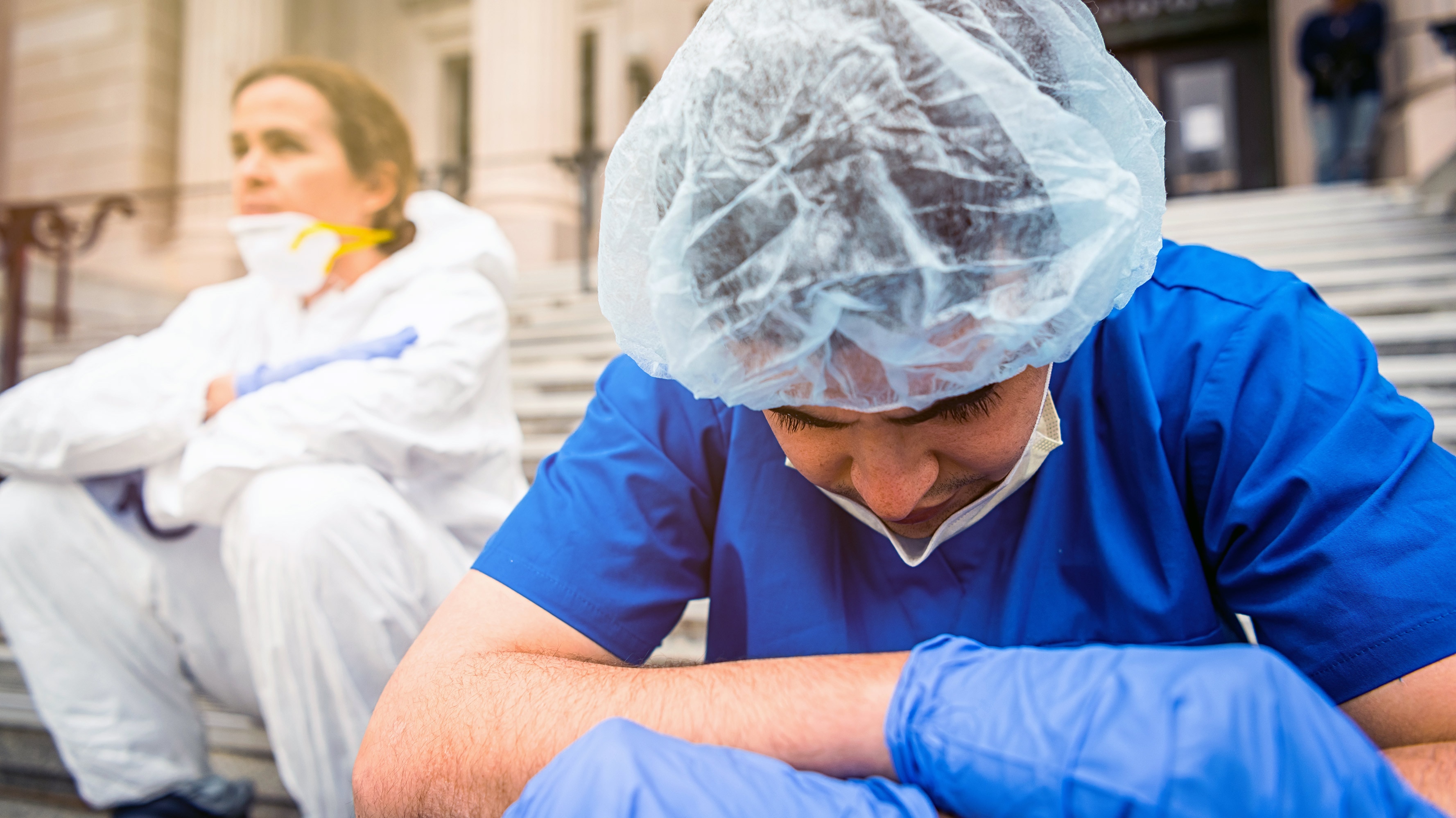 A male doctor, wearing blue scrubs, a face mask, and a hair net, sits on steps outside of a medical facility, looking pensive. A female co-worker sits next to him in the background, looking into the distance.