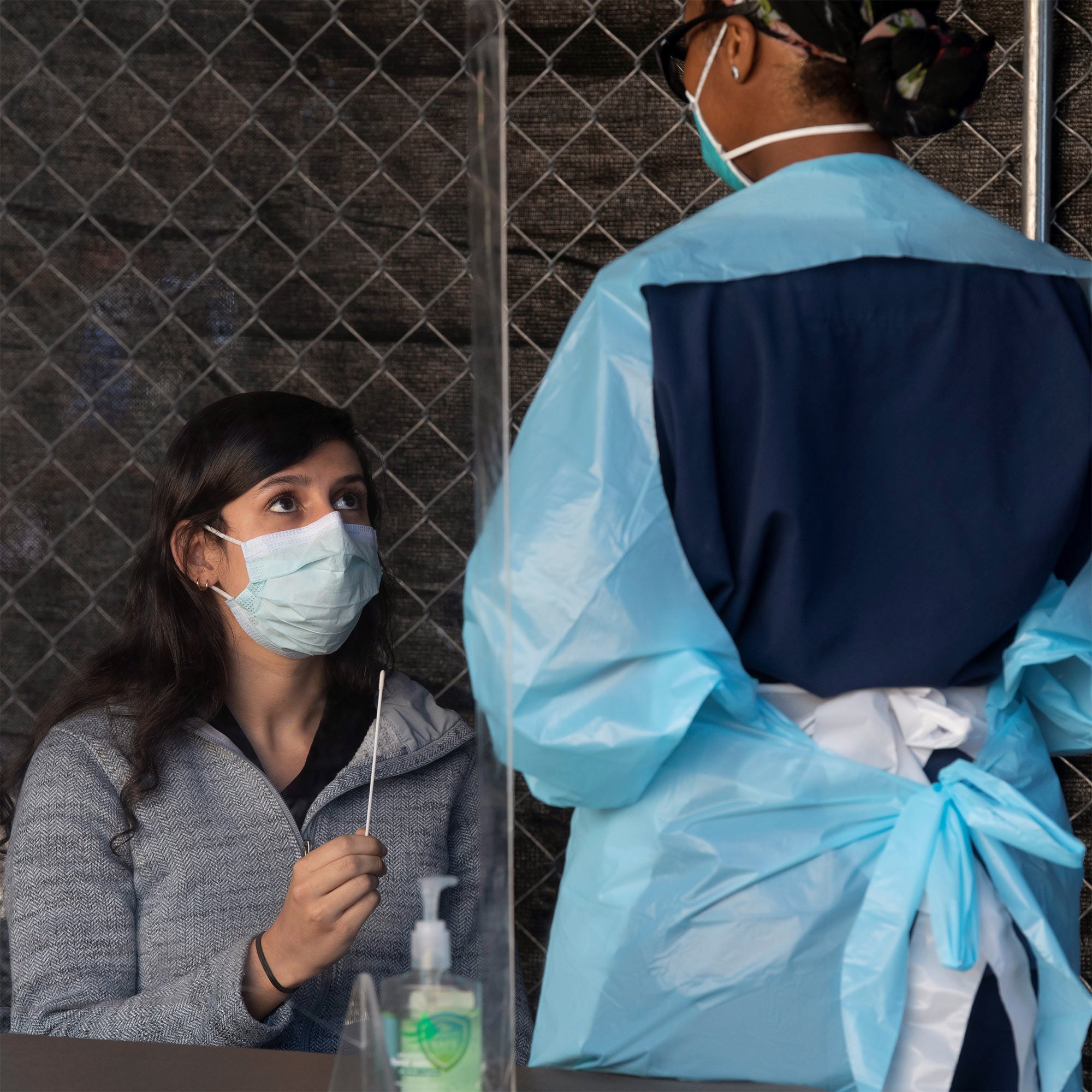 A woman holds a nasal swab as part of a COVID-19 test while a medical professional, (wearing personal protective equipment) advises her on to use the swab to conduct the test.