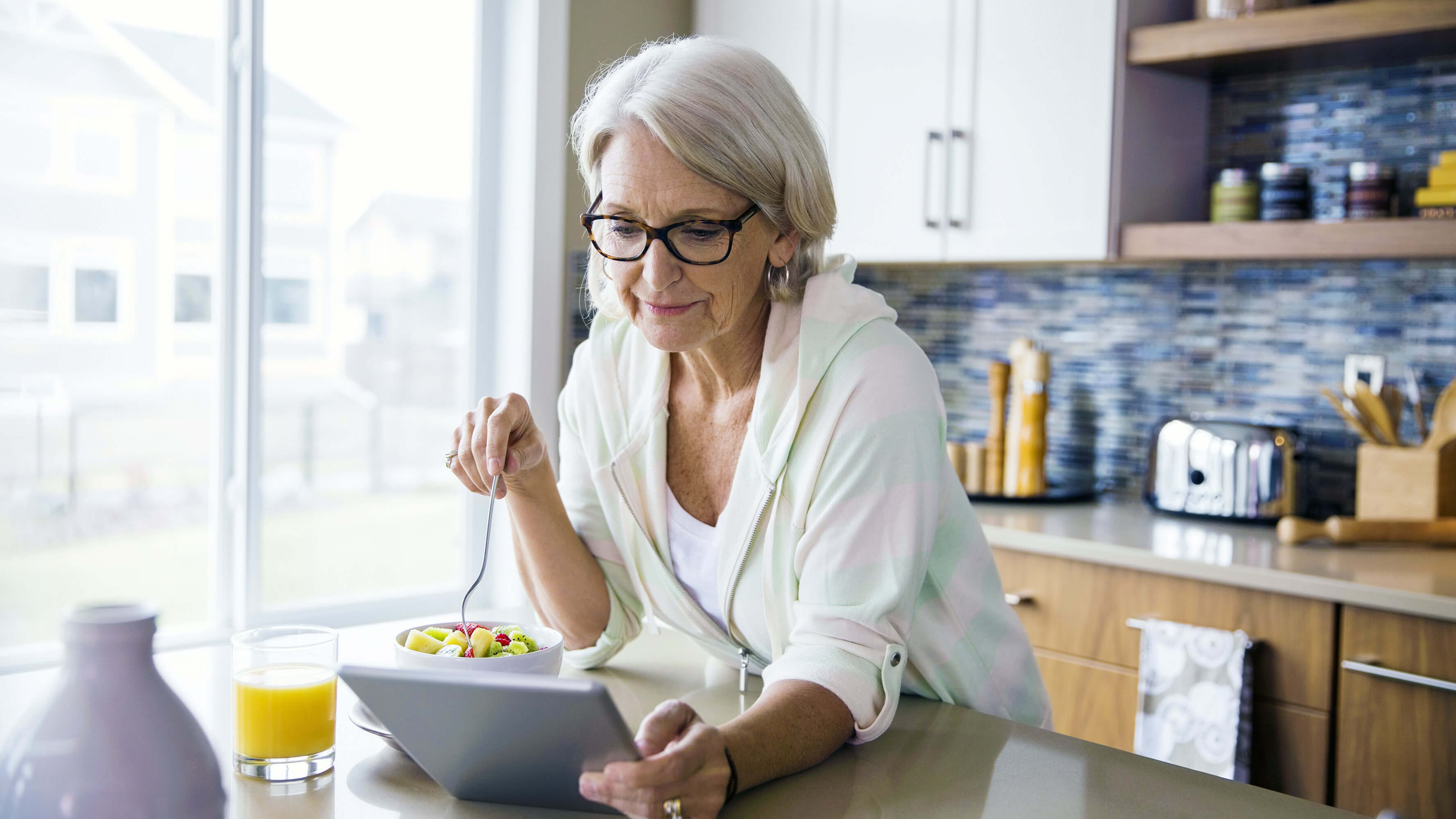 An older female with silvery, long hair eats a bowl of fruit in her kitchen while using a tablet computer to access digital health services.