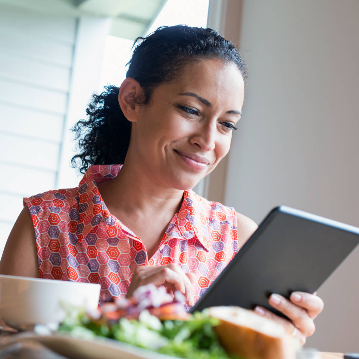 A woman sits at a dining room table eating a salad and a piece of bread while quietly smiling and reading a tablet computer.