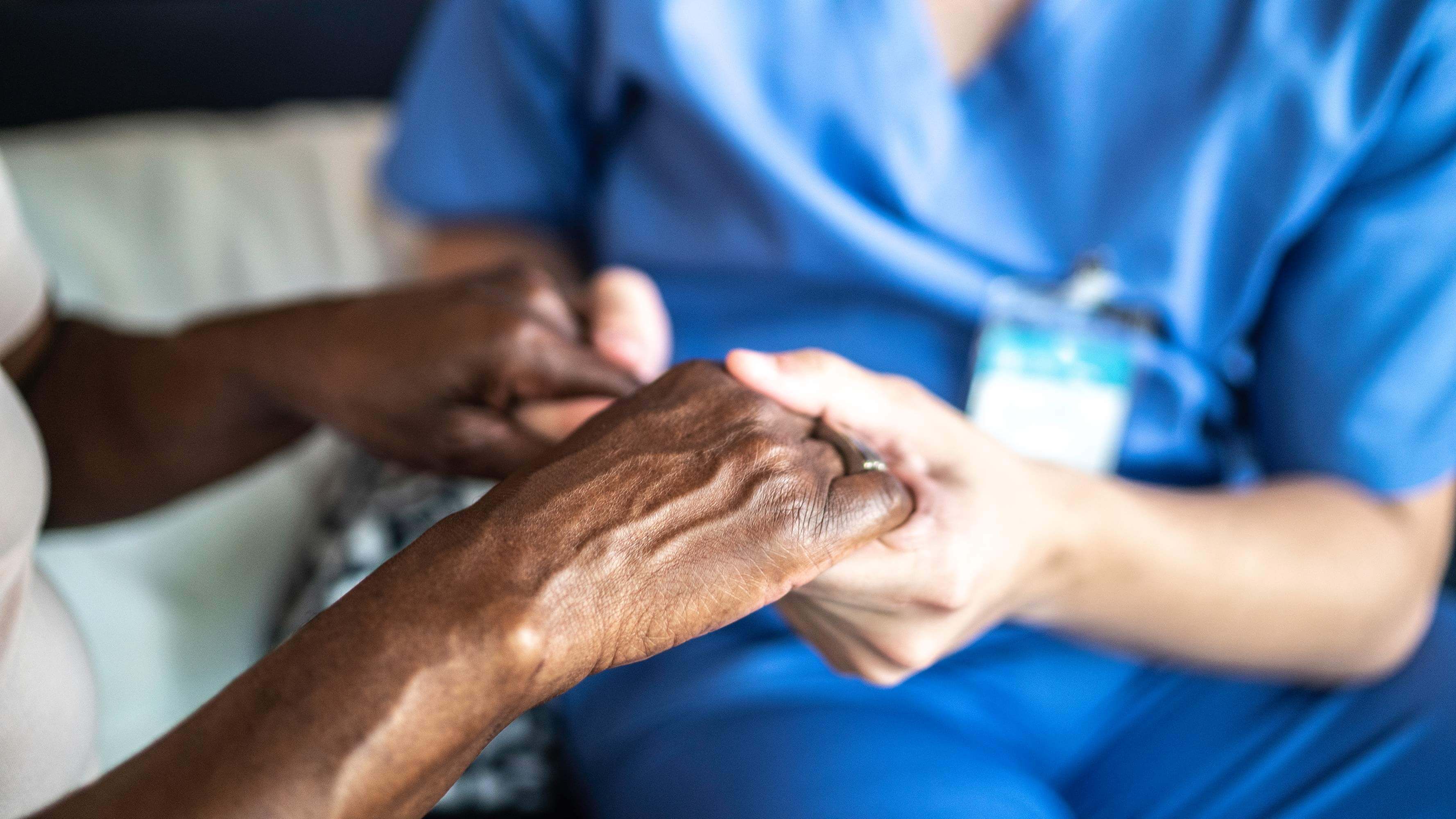 A close-up photo of a nurse, wearing bright blue scrubs, holding the hand of patient.