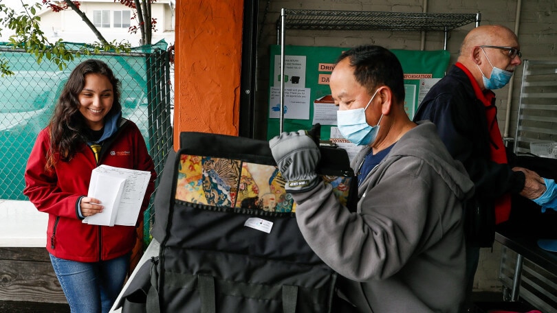A volunteer (wearing a face mask) loads temperature-sensitive food into an insulated bag for distribution. A woman in a red coat observes him loading the insulated bag.