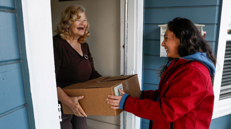 A female volunteer, wearing a red coat, delivers a box containing food donations to female senior citizens, who is smiling while standing in the doorway of her house.