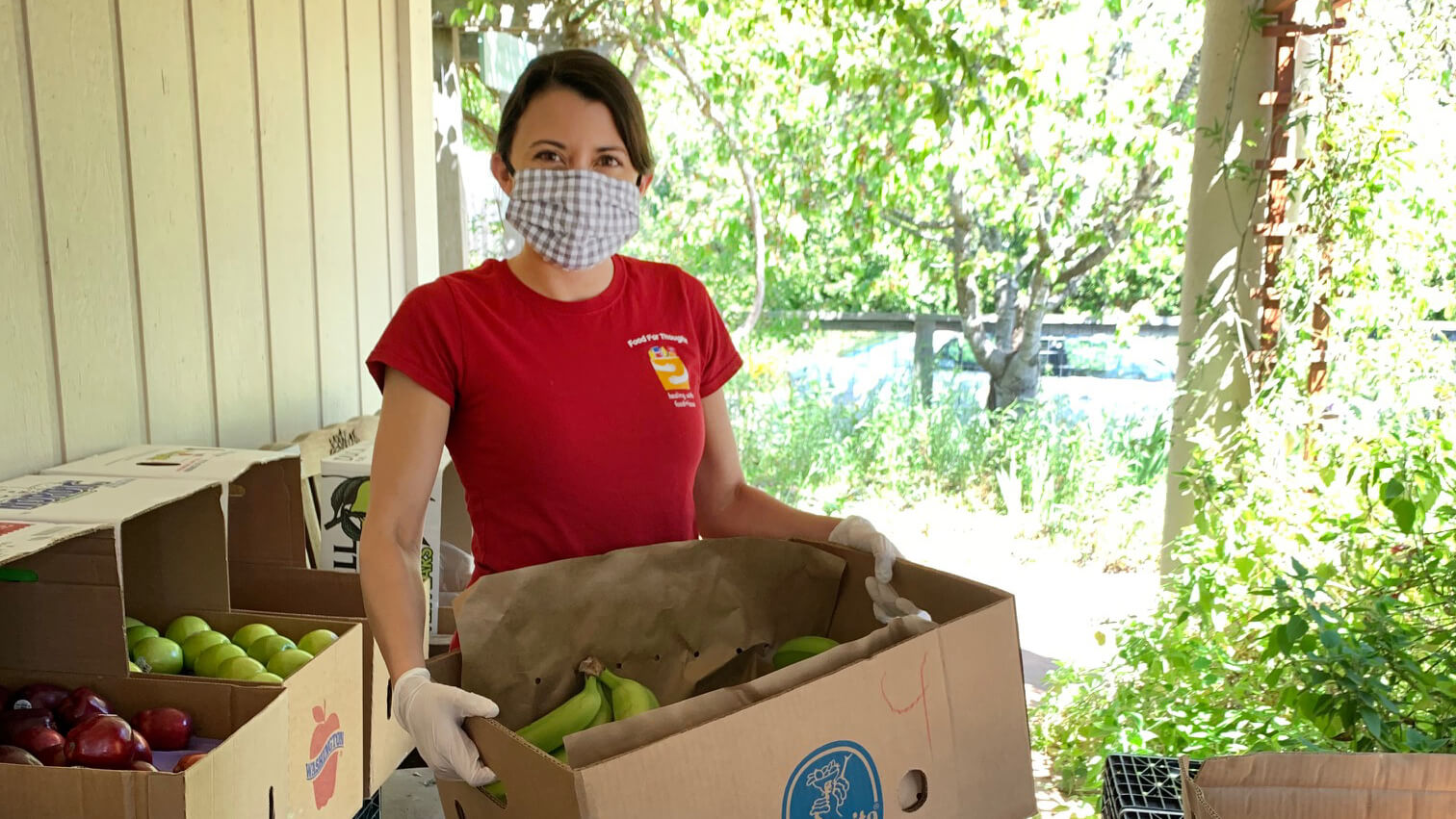A woman, wearing a face mask and gloves, handles produce in cardboard boxes.