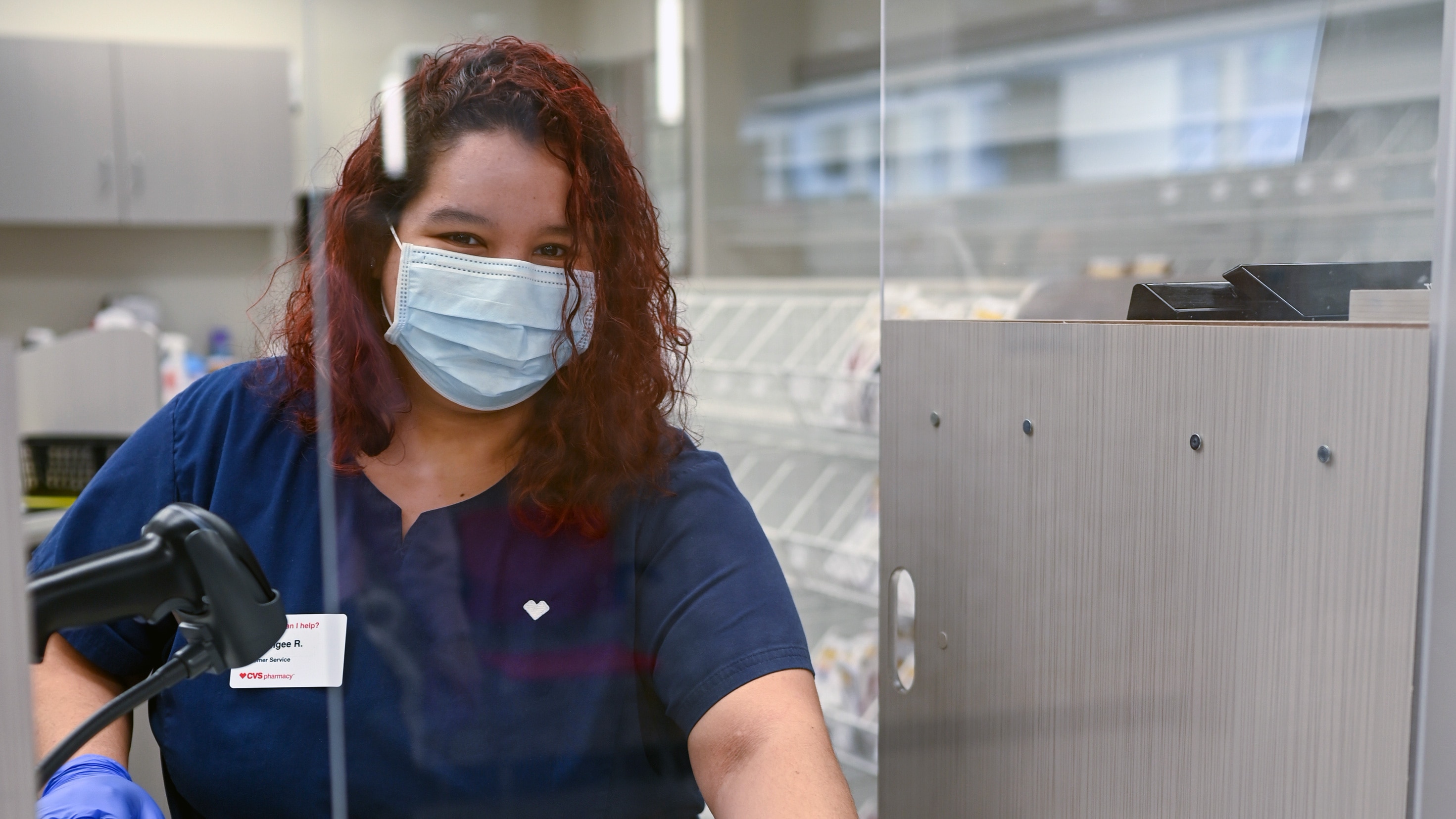 A CVS Pharmacy technician prepares prescriptions while wearing personal protective equipment (PPE) behind a plexiglass screen.