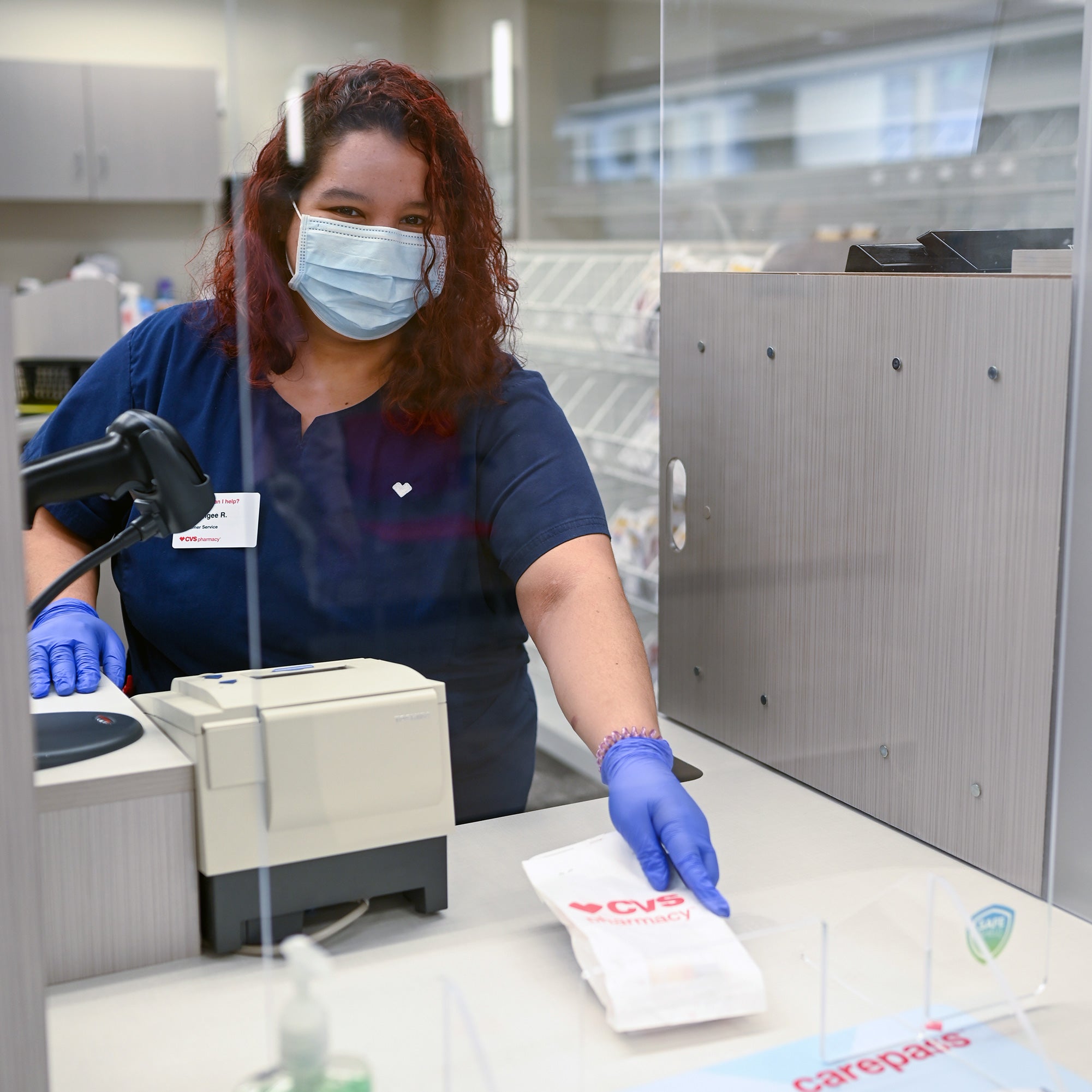 A CVS Pharmacy technician prepares prescriptions while wearing personal protective equipment (PPE) behind a plexiglass screen.