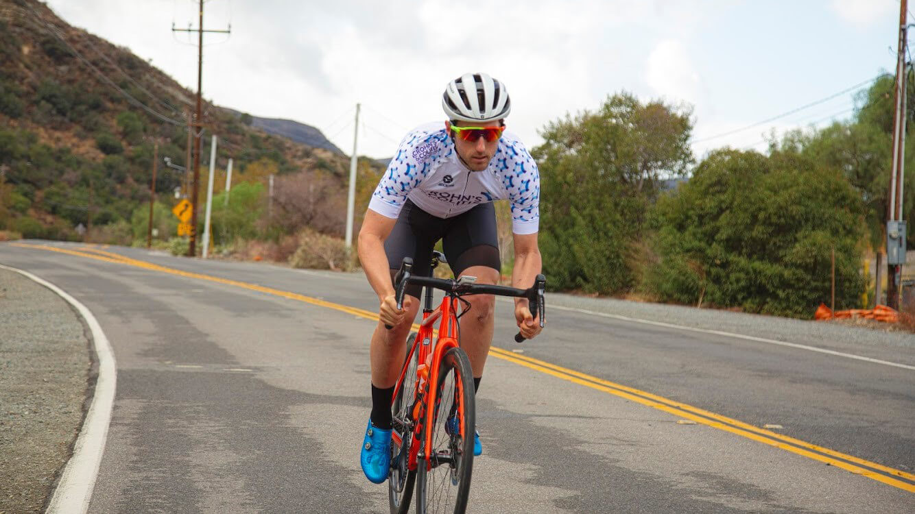 Photo of Cory Greenberg cycling on a road with a mountain in the background.