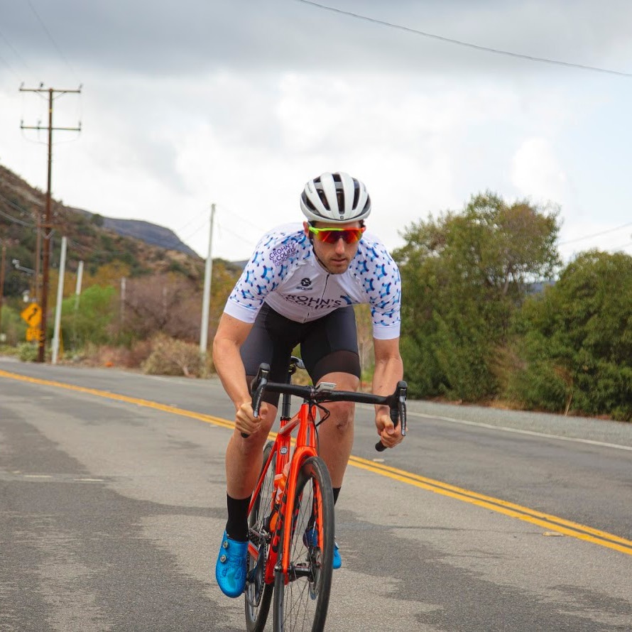 Photo of Cory Greenberg cycling on a road with mountains in the background.