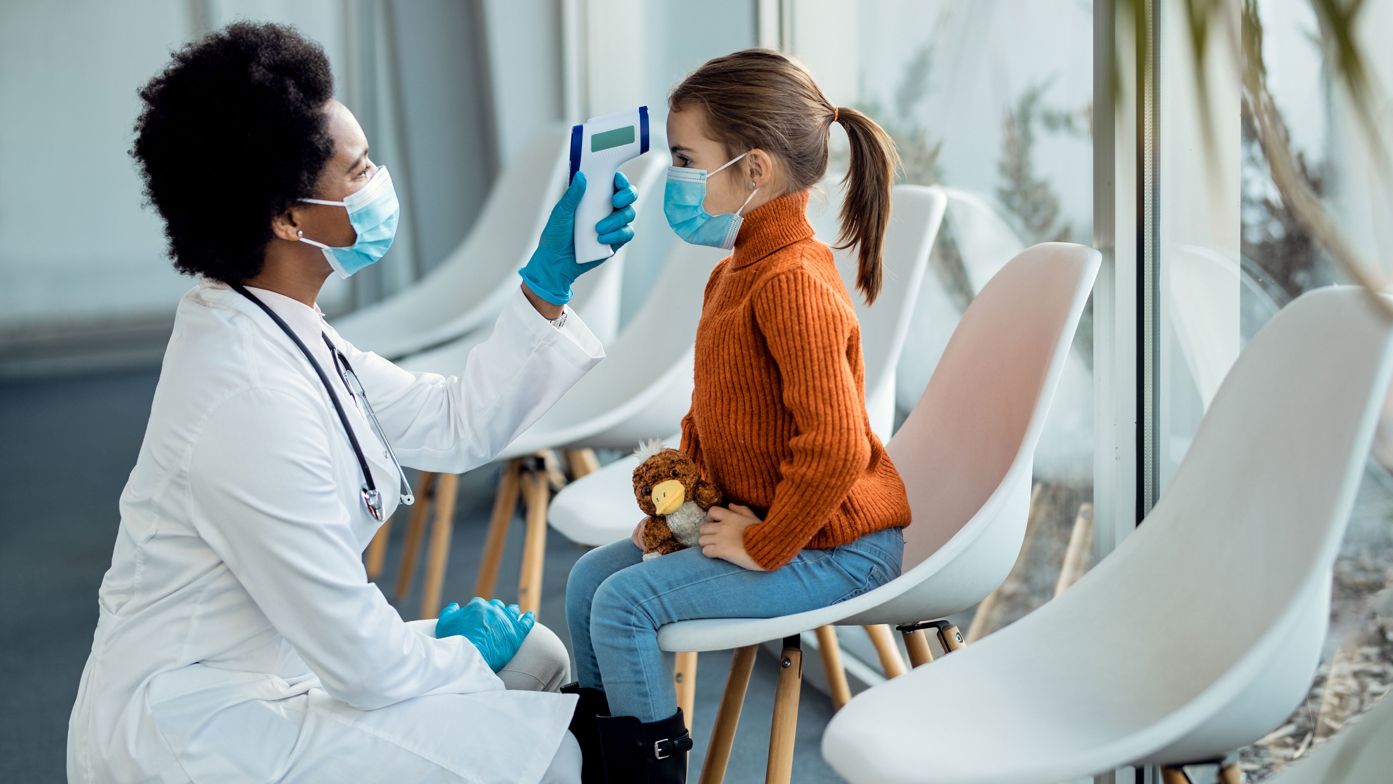 A doctor kneeling in front of a small girl sitting on a chair taking her temperature