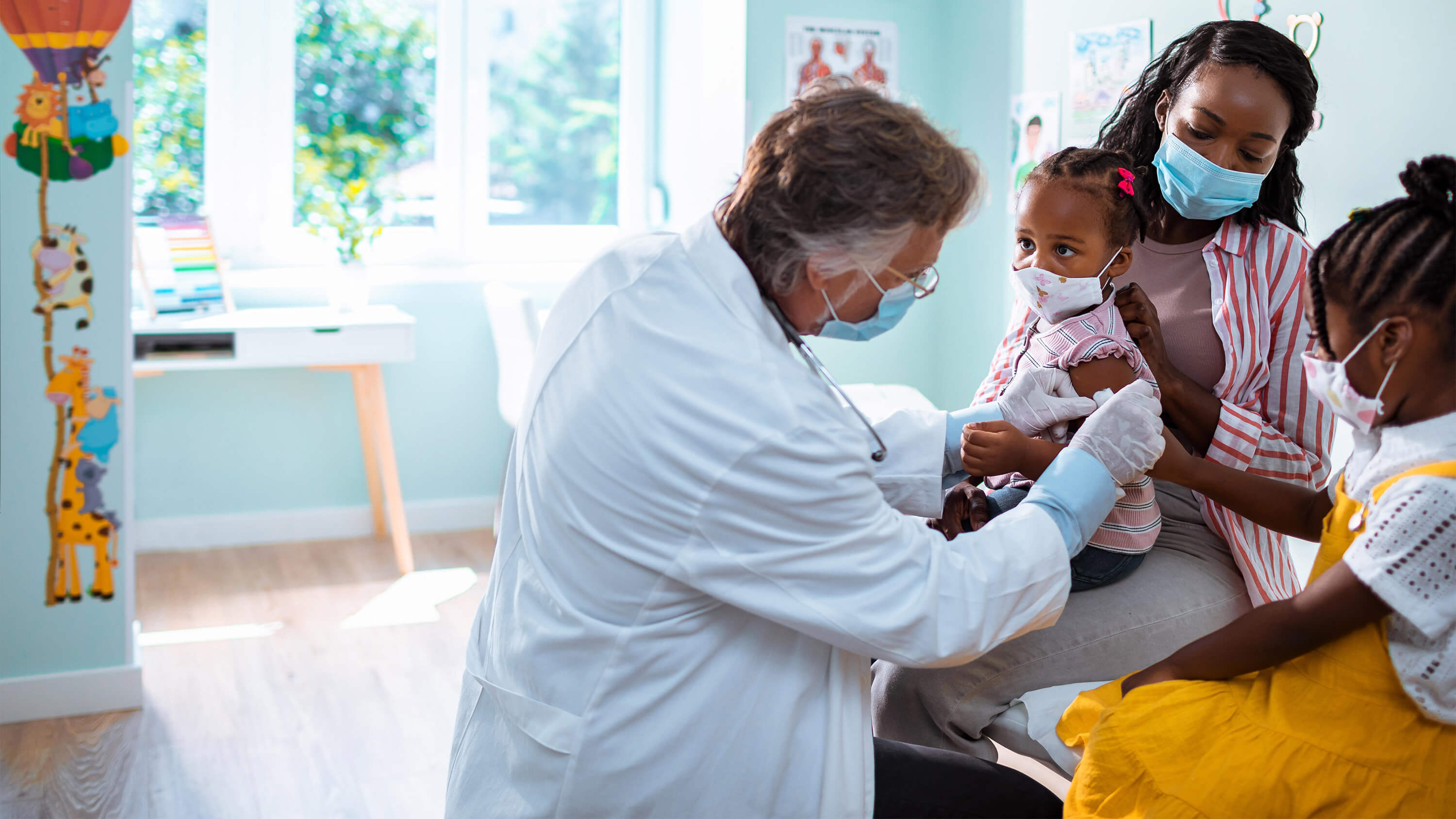 A physician administers a vaccine to a young girl, while her sister and mother look on; all wearing face masks.