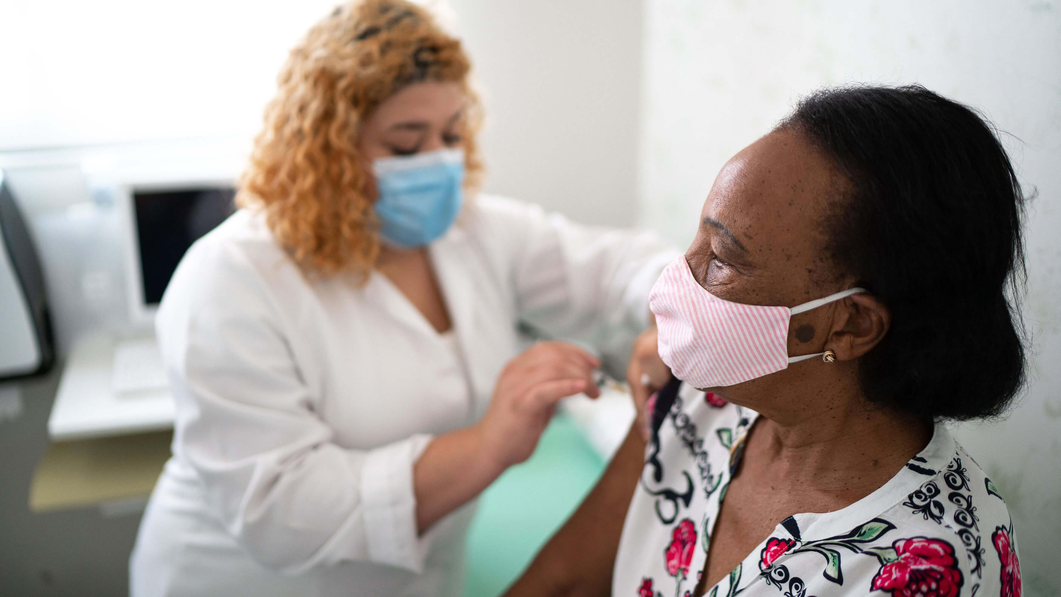 A woman, wearing a face mask, receives a vaccination in an examination room from a nurse practitioner, also wearing a face mask.