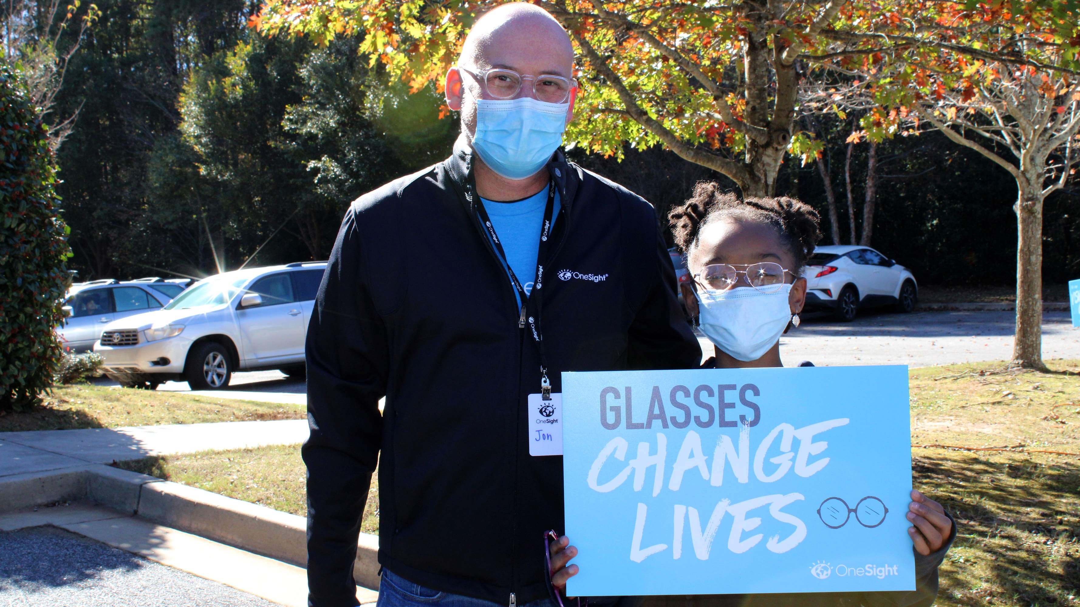 Jon, a OneSight team member, stands with a young girl wearing glasses, both wearing face masks, with a sign that reads, “Glasses change lives”.