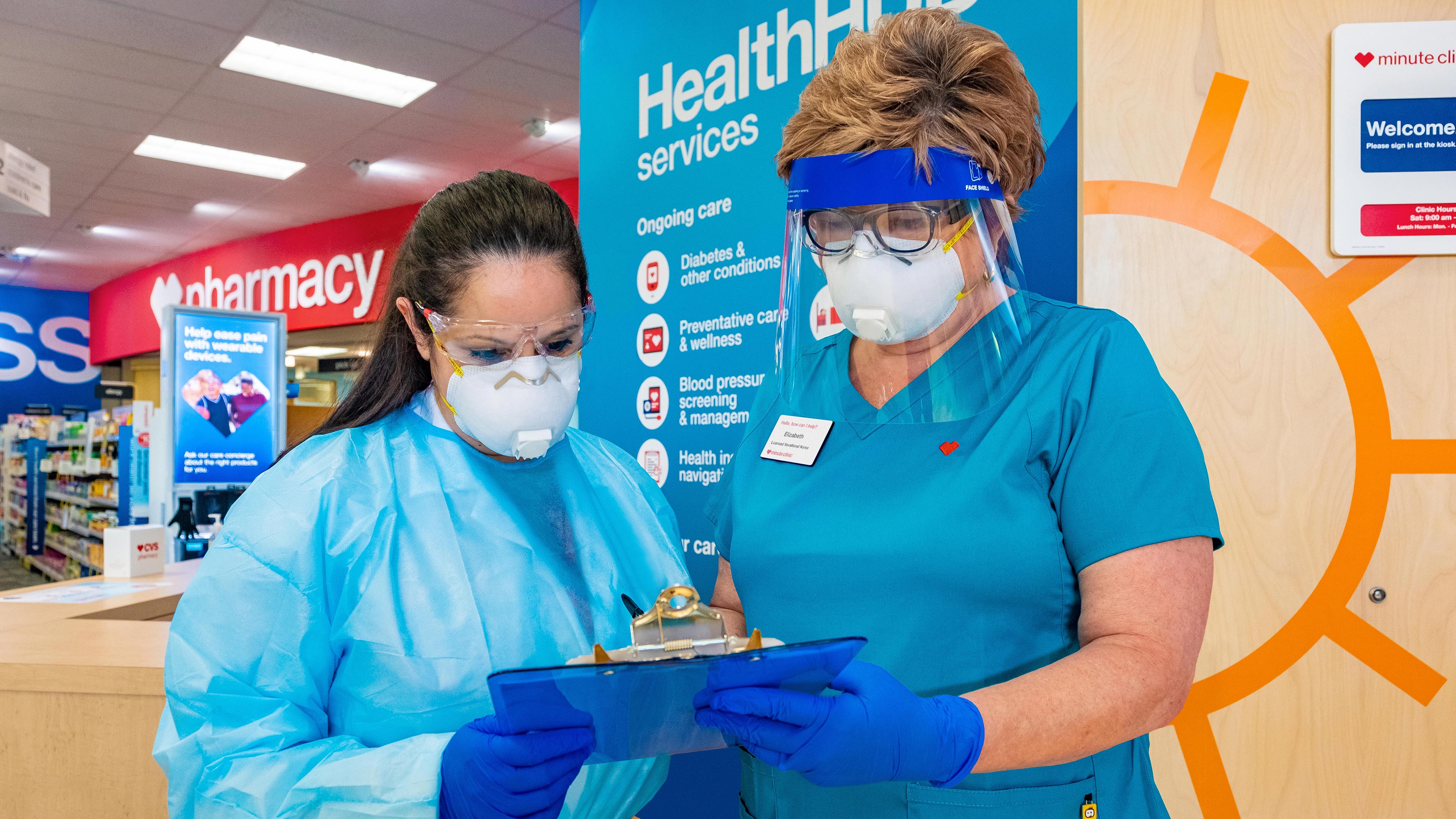 Two CVS Pharmacy technicians, wearing personal protective equipment (PPE), go over paperwork in a HealthHUB location.