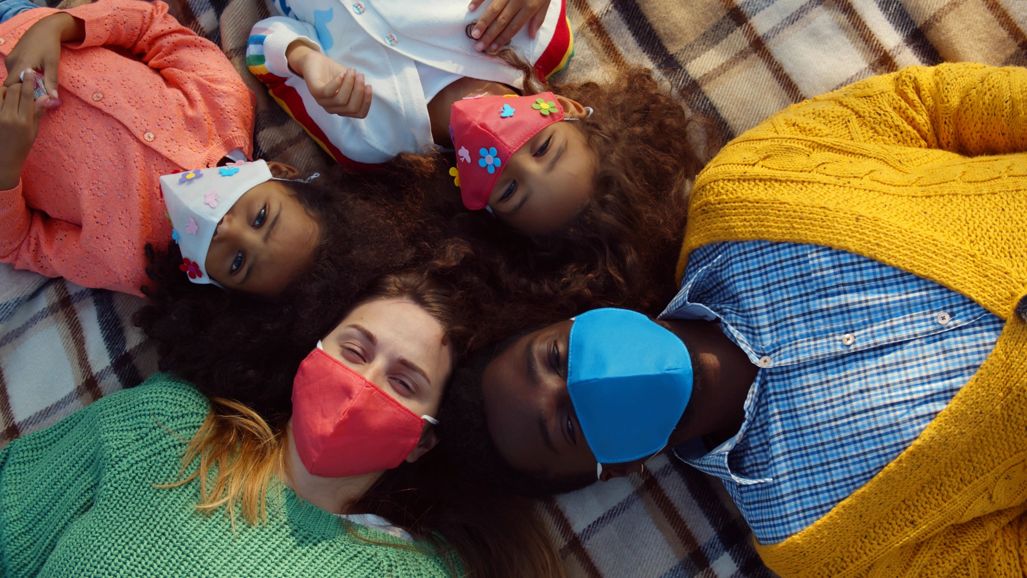 A family of four lying in a circle on the floor wearing masks looking up at the camera
