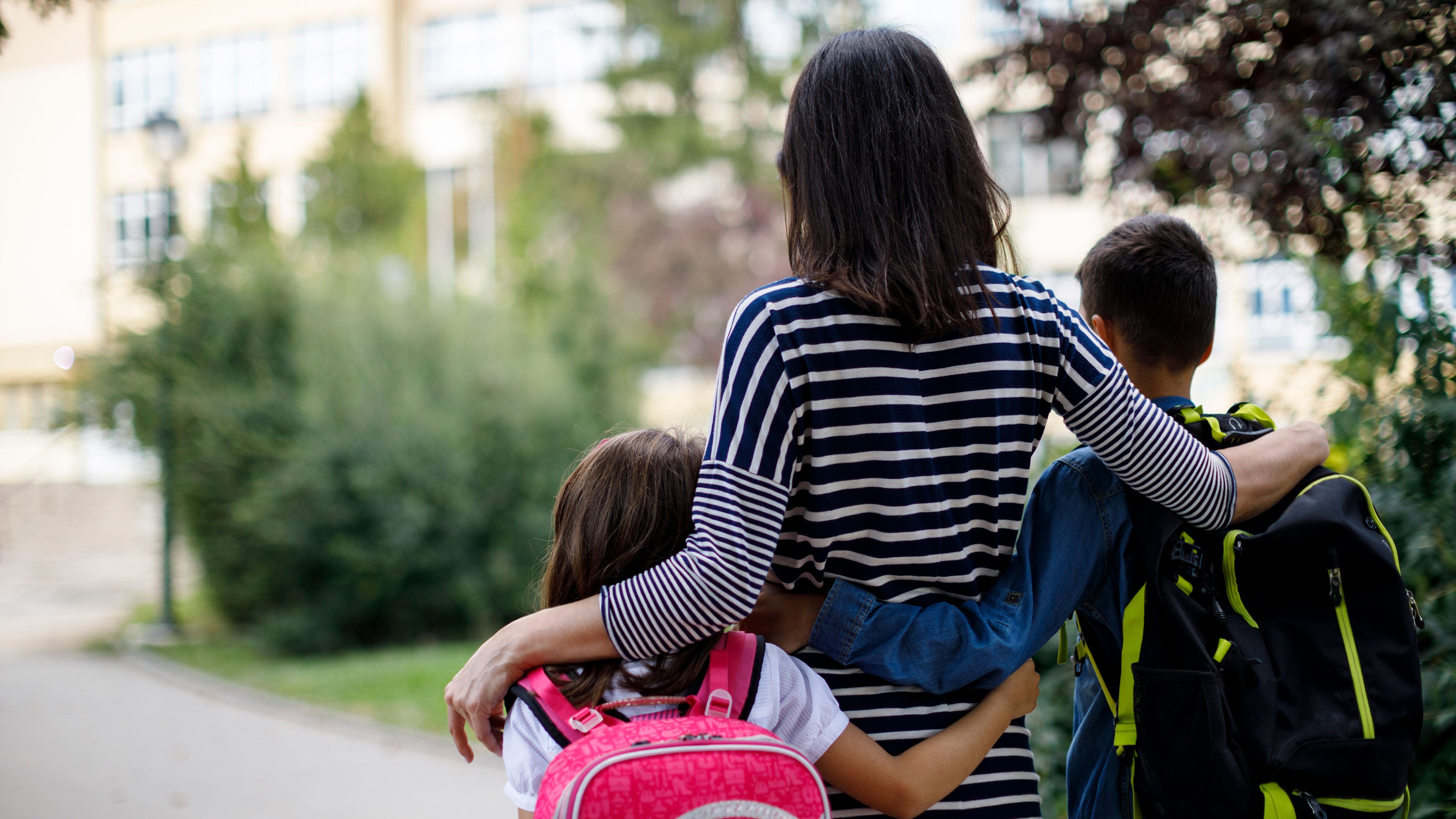 A young mother walking with her arms around her two children on their way to school. 