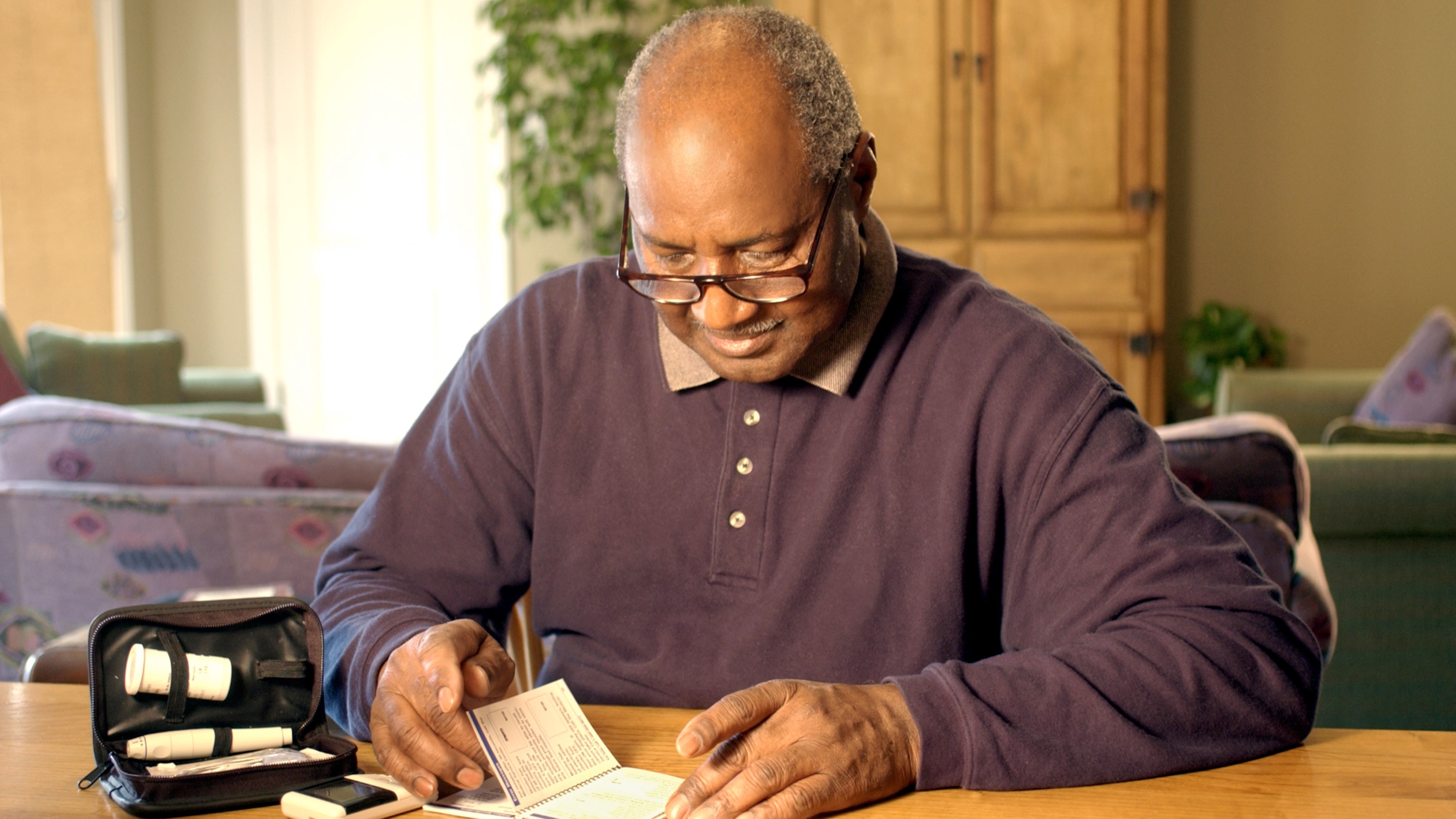 An older black man seated at a table reading manual for blood test kit.
