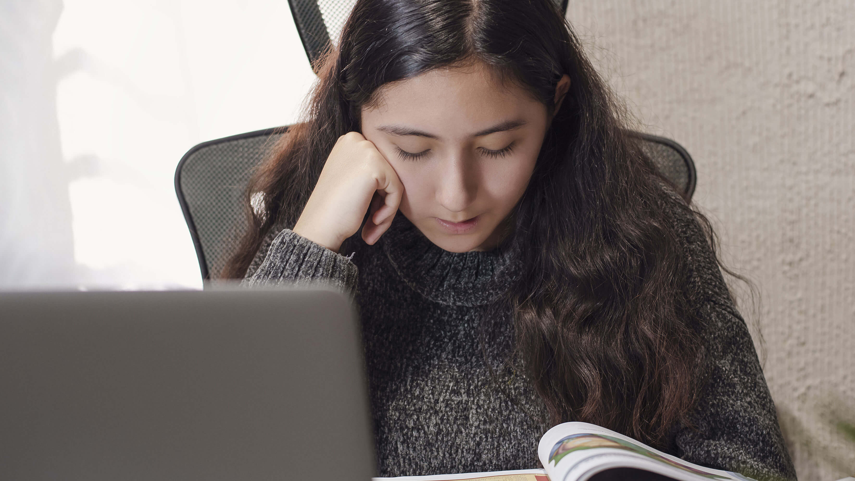 A girl in a grey sweater reads a book in front of her laptop while resting her cheek on her hand.