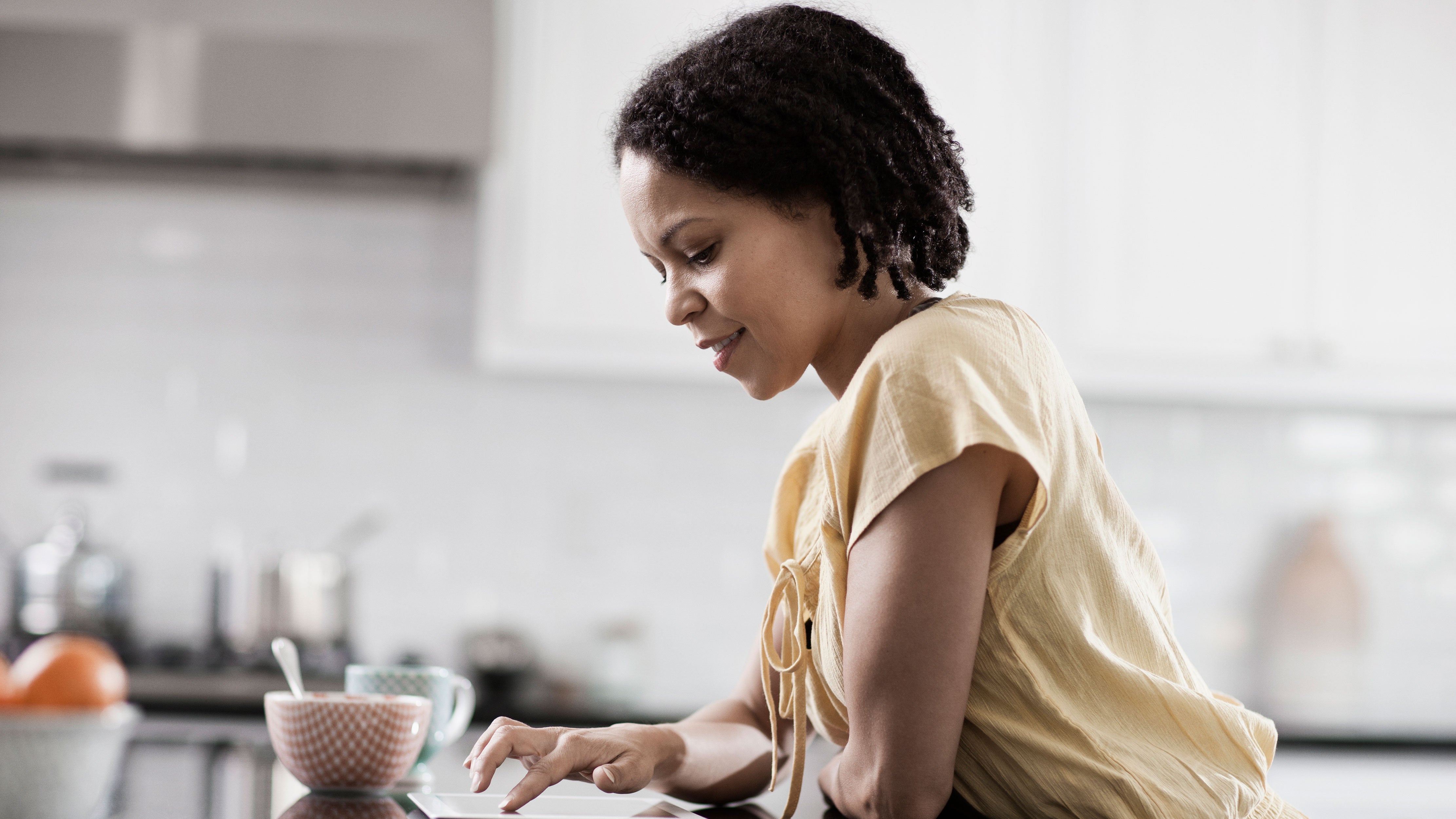 A woman sits at her kitchen counter with her breakfast to scroll on her cell phone.
