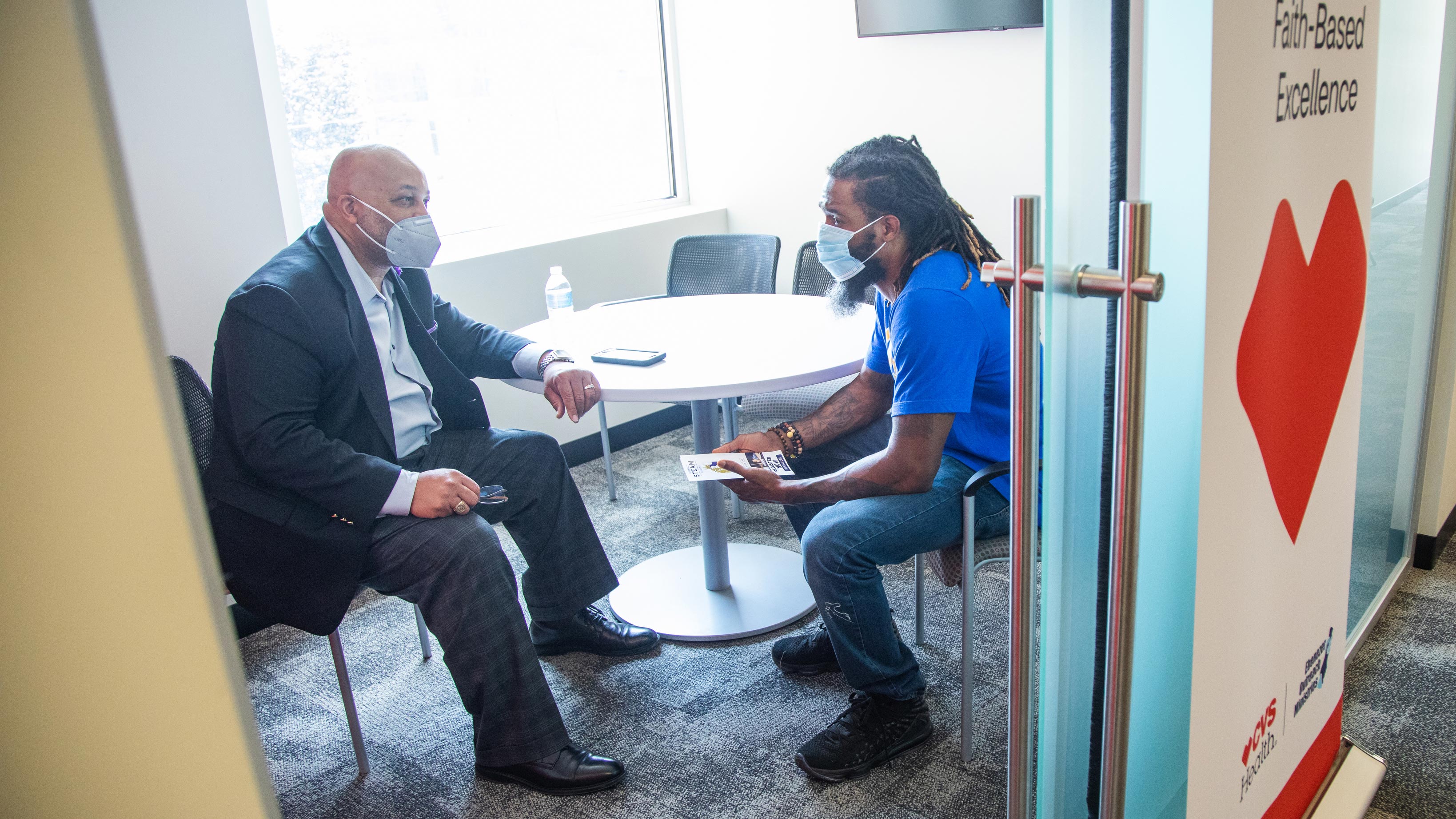 Two masked men sit in a conference room to discuss work training.