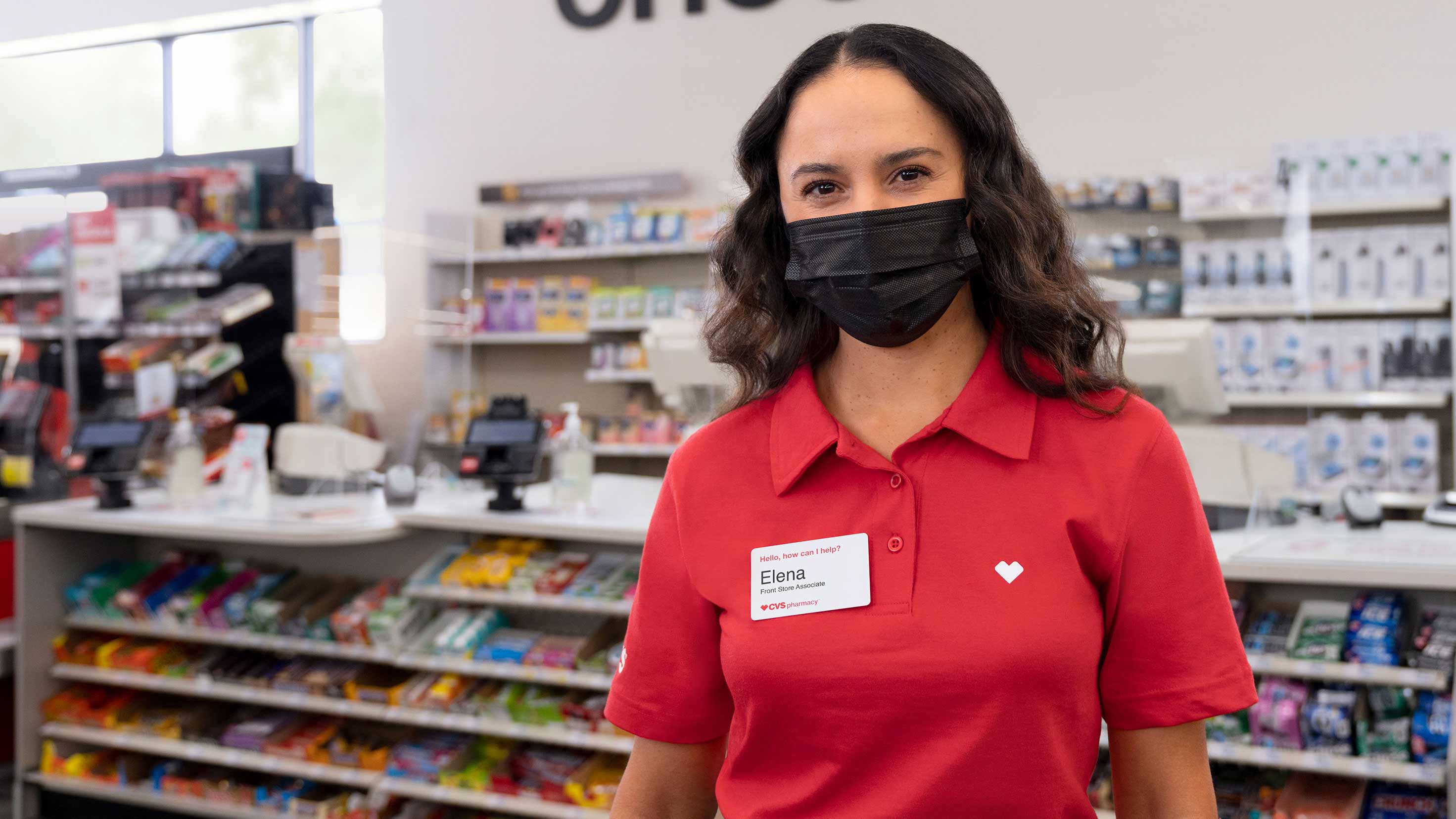A CVS Pharmacy colleague, wearing a face mask, stands in front of a store checkout area.