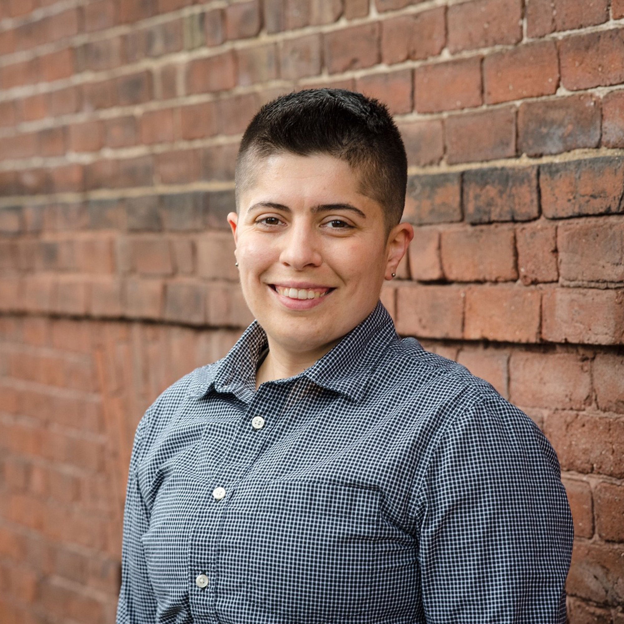 Jess Coppola wears a blue, checkered button-down and stands in front of a brick wall to smile for a picture.