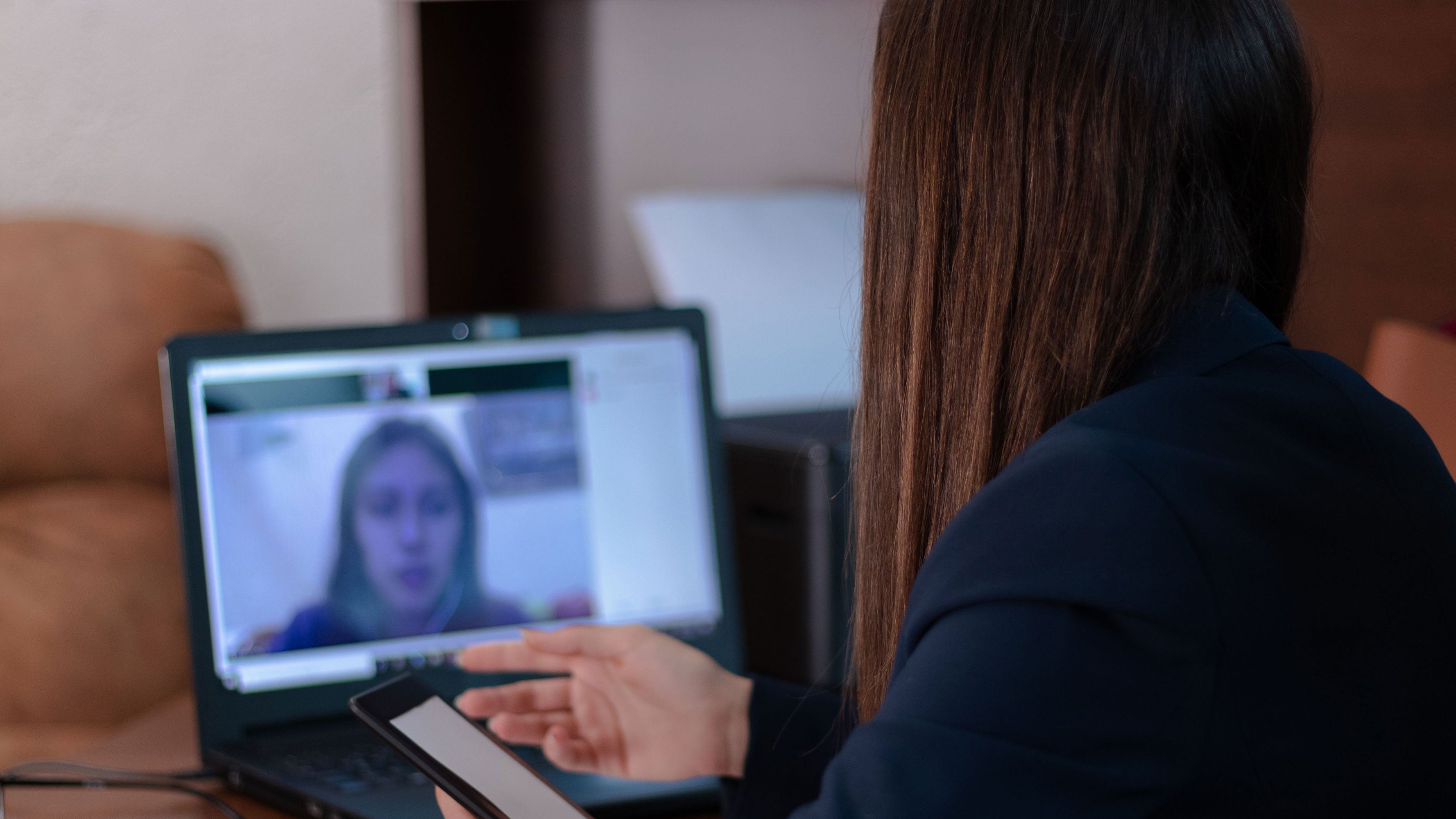 A woman uses her smartphone while she sits at her laptop to videochat.