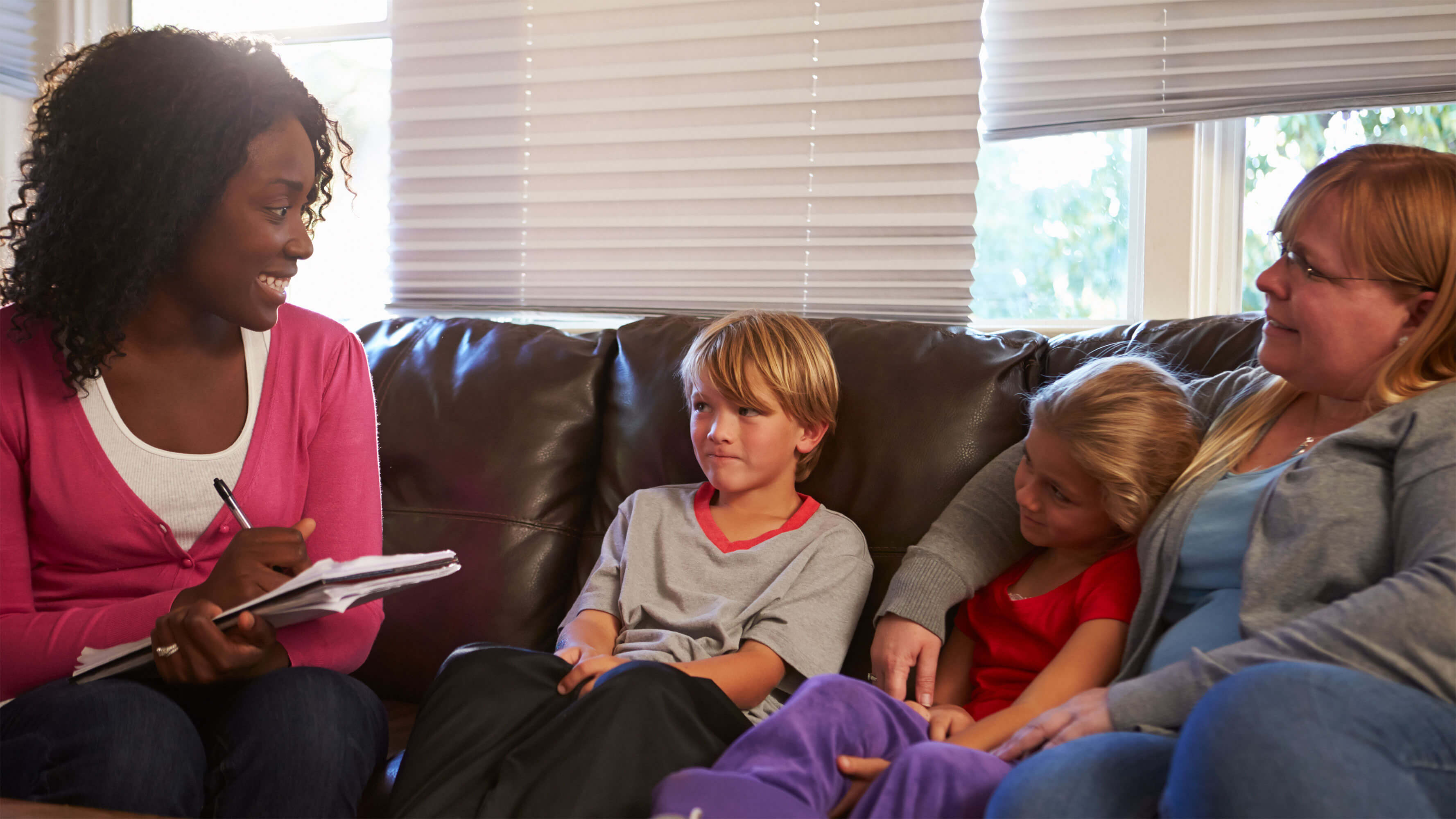 A woman wearing a pink sweater takes notes in a notebook while chatting with a mother and her two young children.