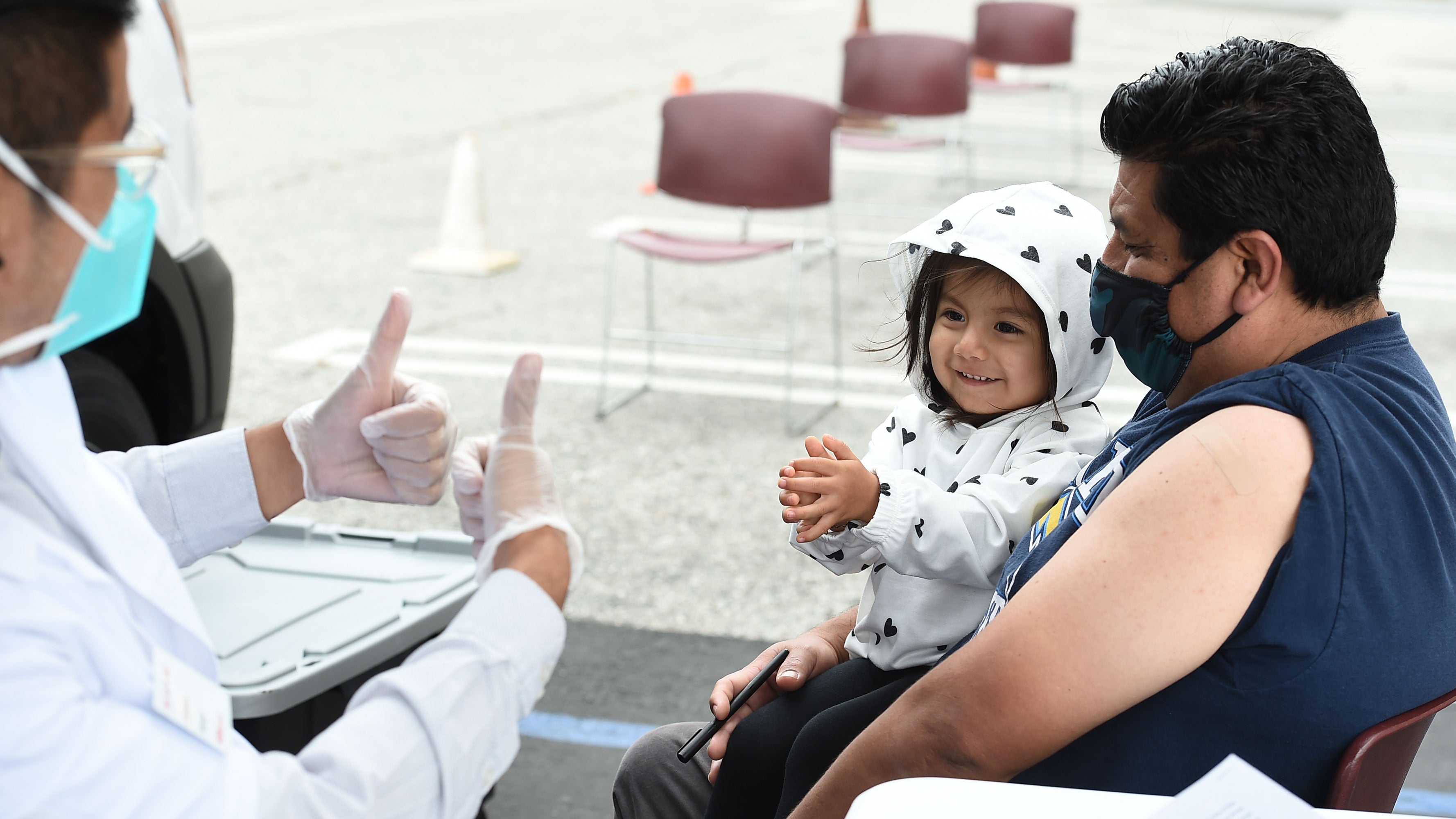 A medical worker, wearing PPE, offers two thumbs up to a young girl sitting in her father&#039;s lap.