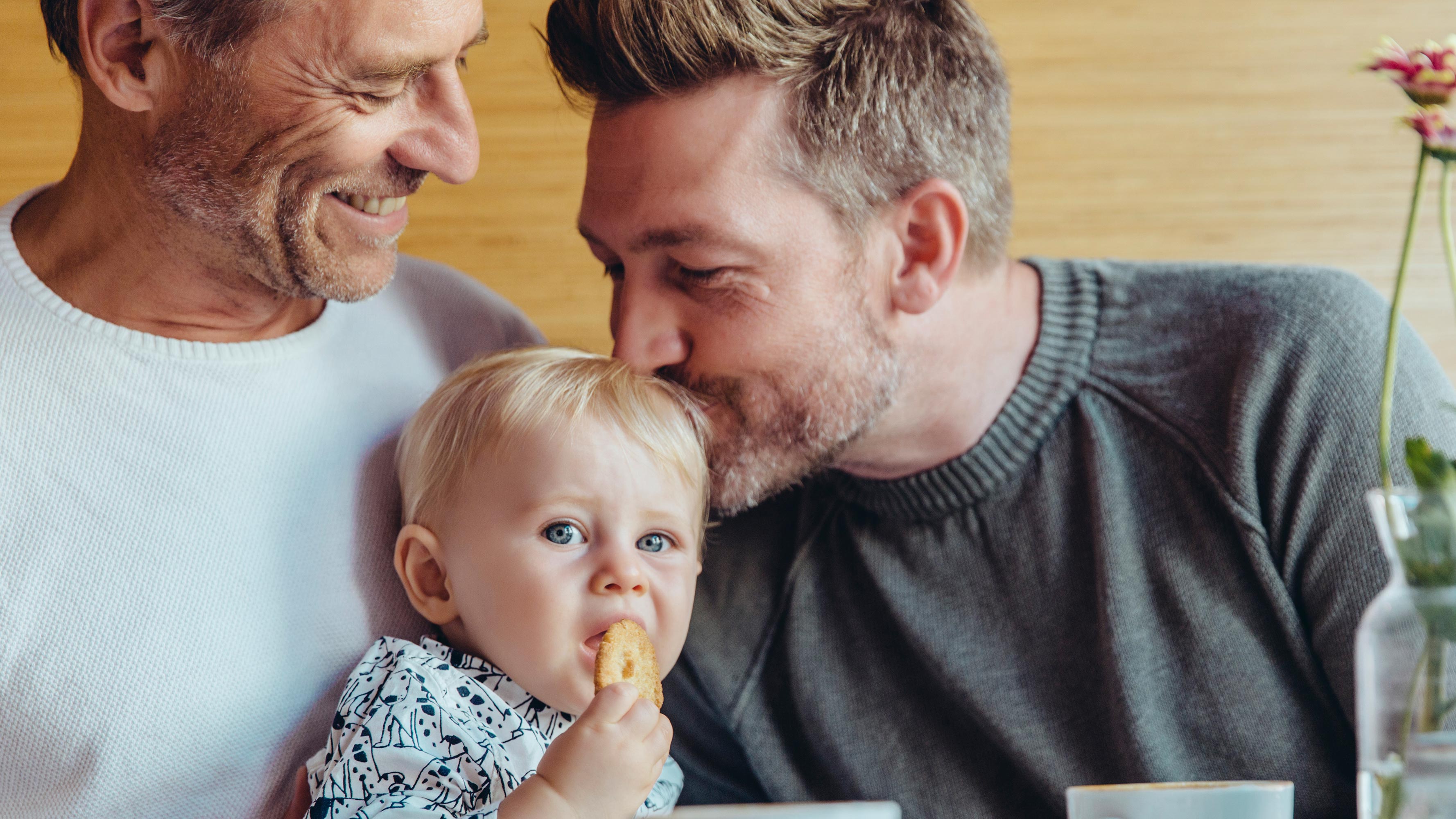 Two fathers sit at a table with their young daughter. One kisses the side of her head while the other smiles at his partner.