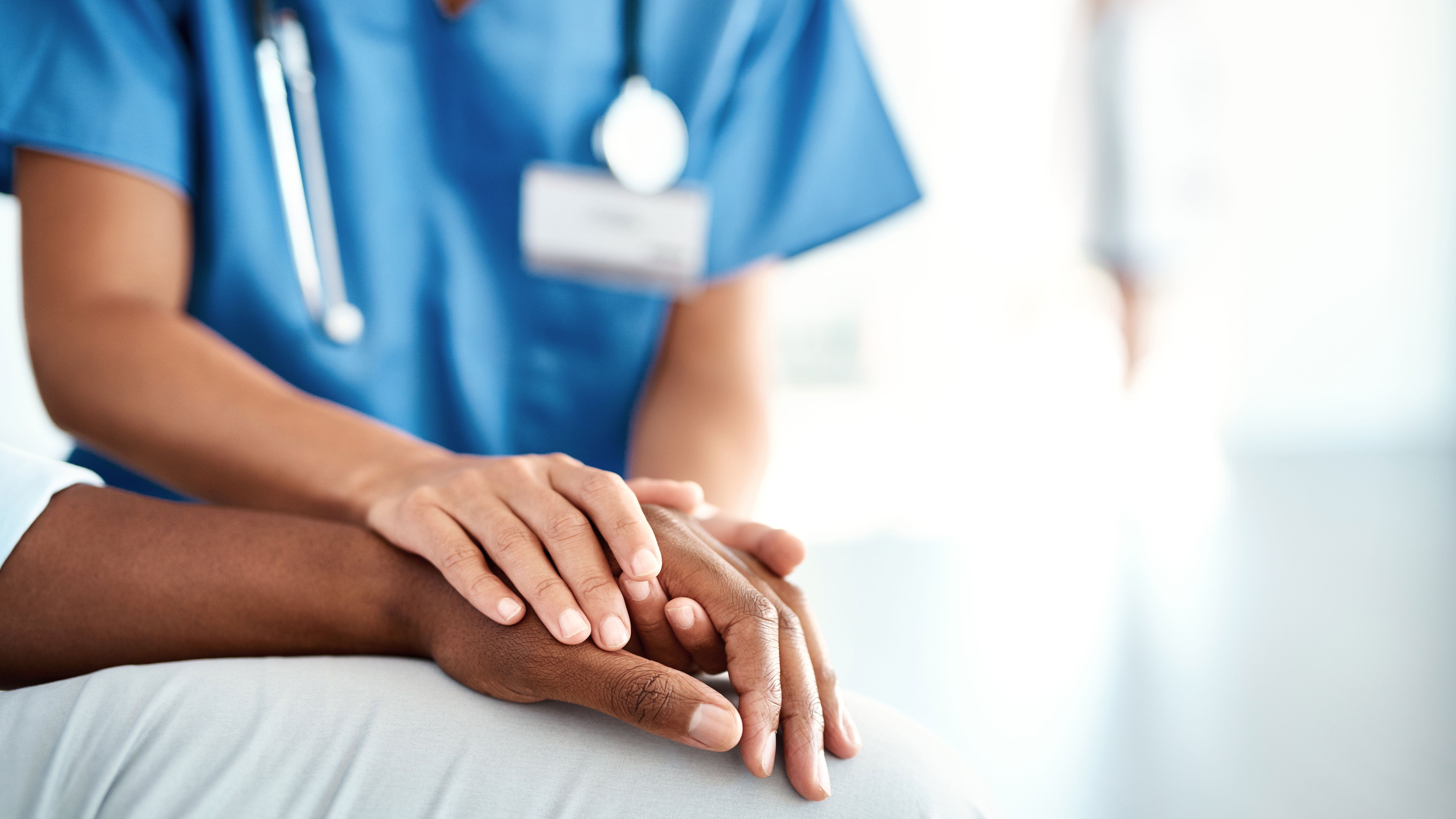A doctor, wearing blue scrubs and a stethoscope around their neck, places their hands on a patient&#039;s hand in comfort.