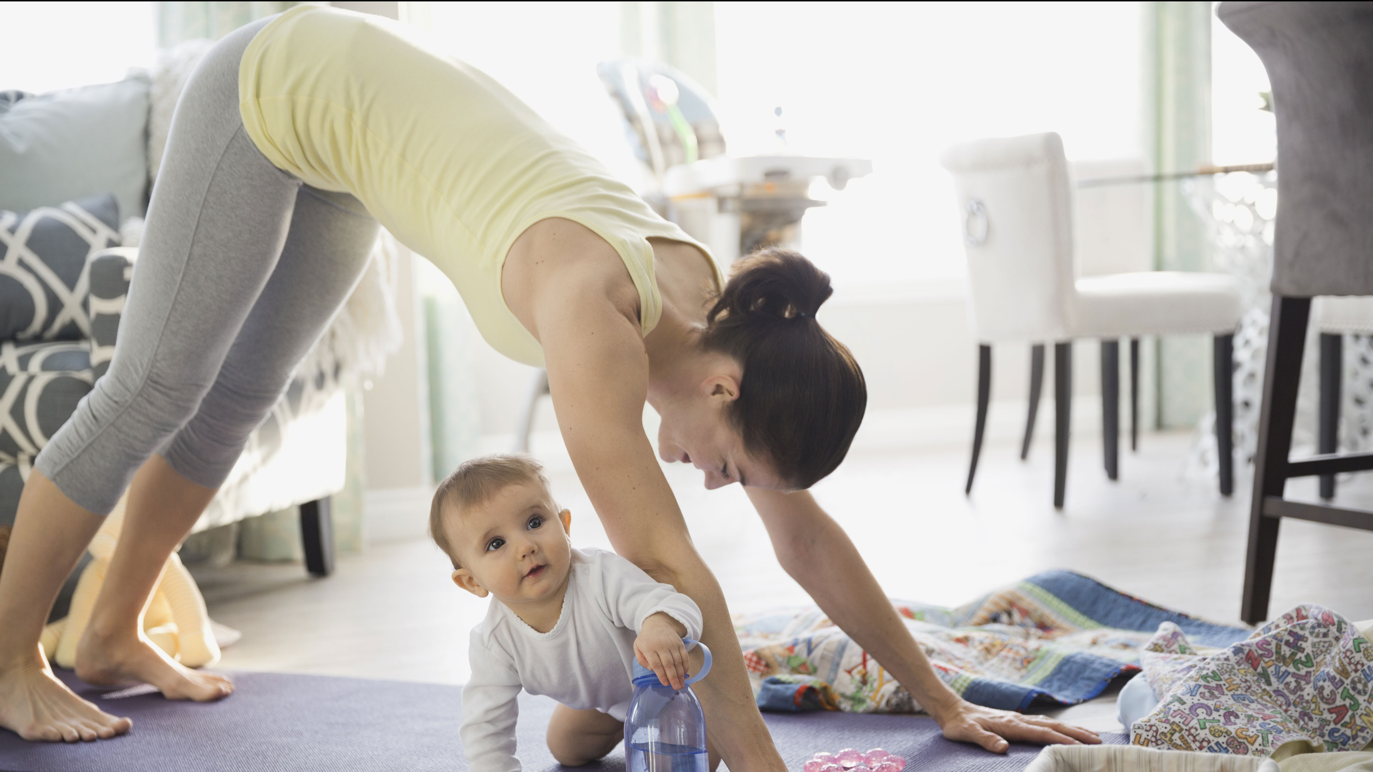 A young child crawls across their mother&#039;s yoga mat as their mother attempts to fold into downward dog pose.