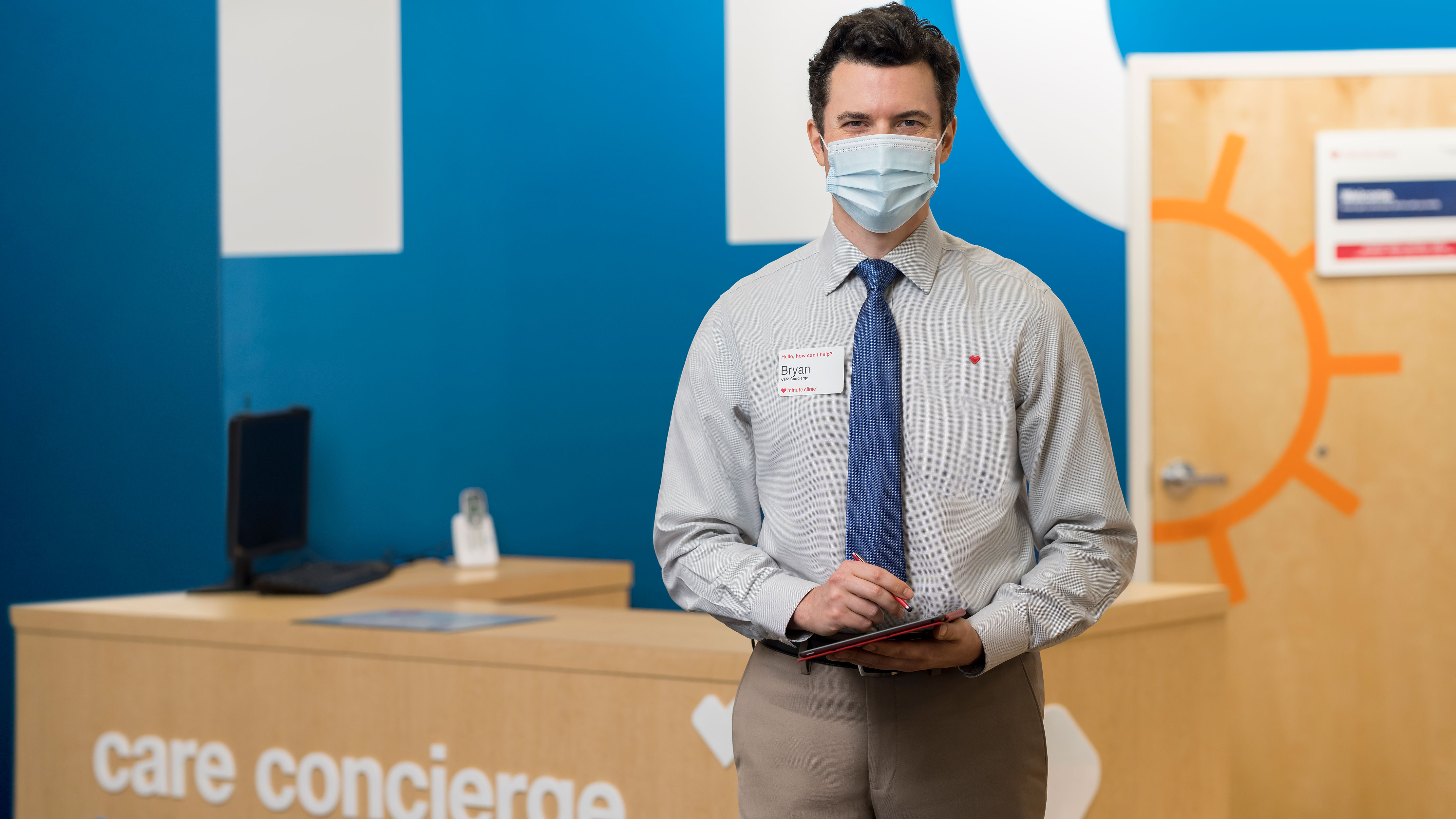 A masked Care concierge stands ready to greet guests at the check-in station for a HealthHUB inside a CVS MinuteClinic
