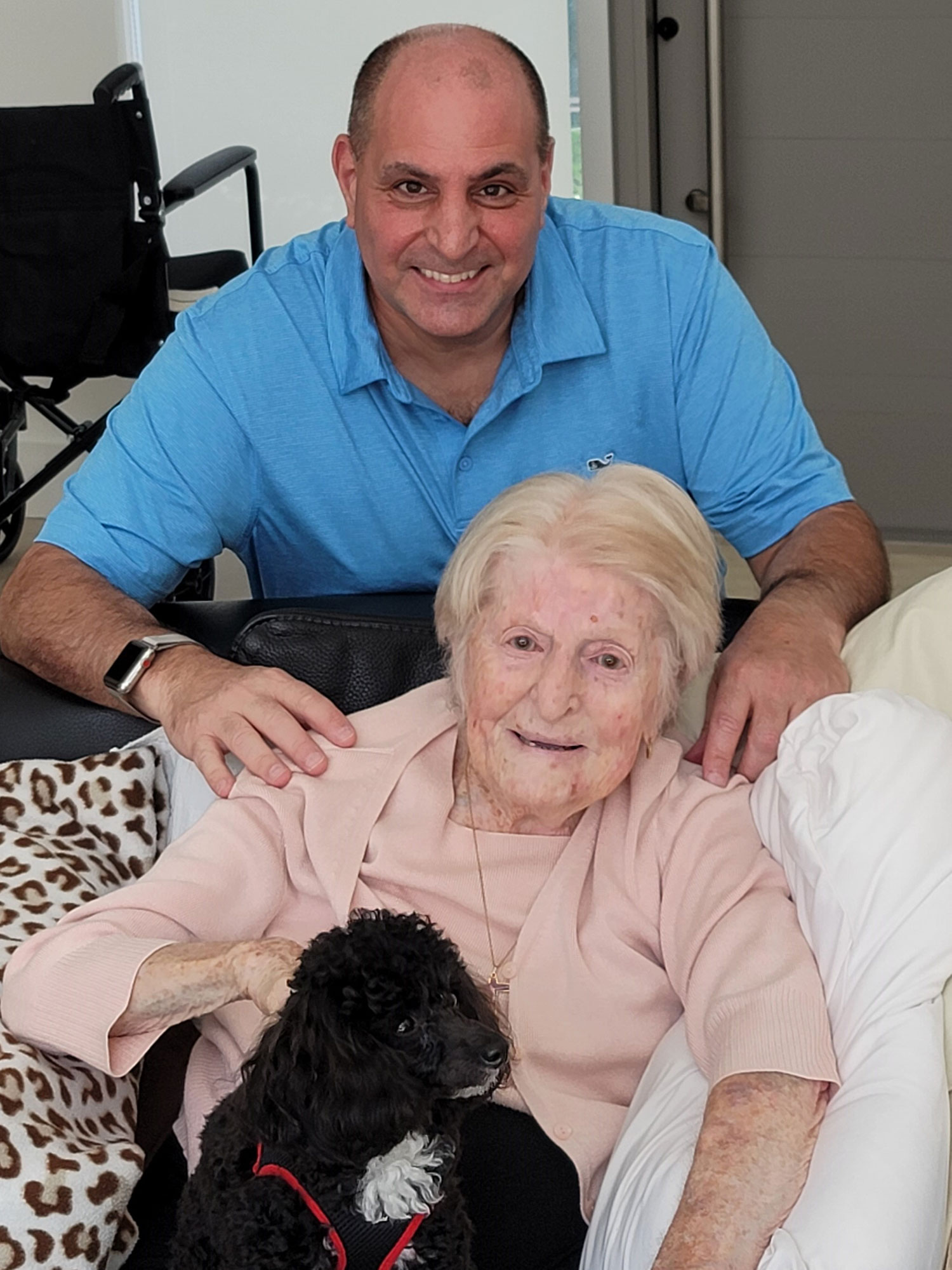 An elderly woman sits with her black dog on her lap while a caretaker smiles.