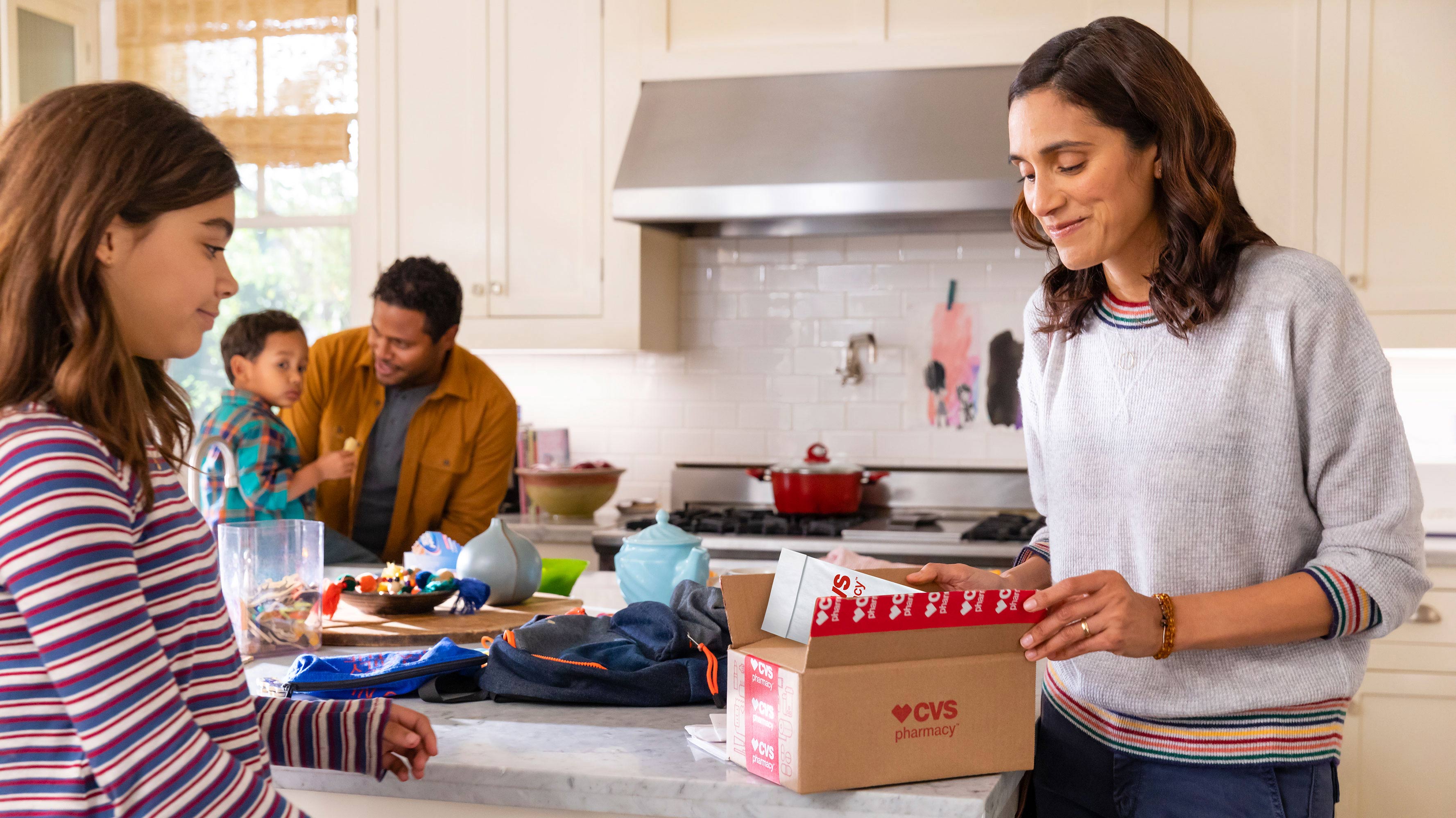 A female, surrounded by her family in their kitchen, opens a delivery box from CVS Pharmacy.