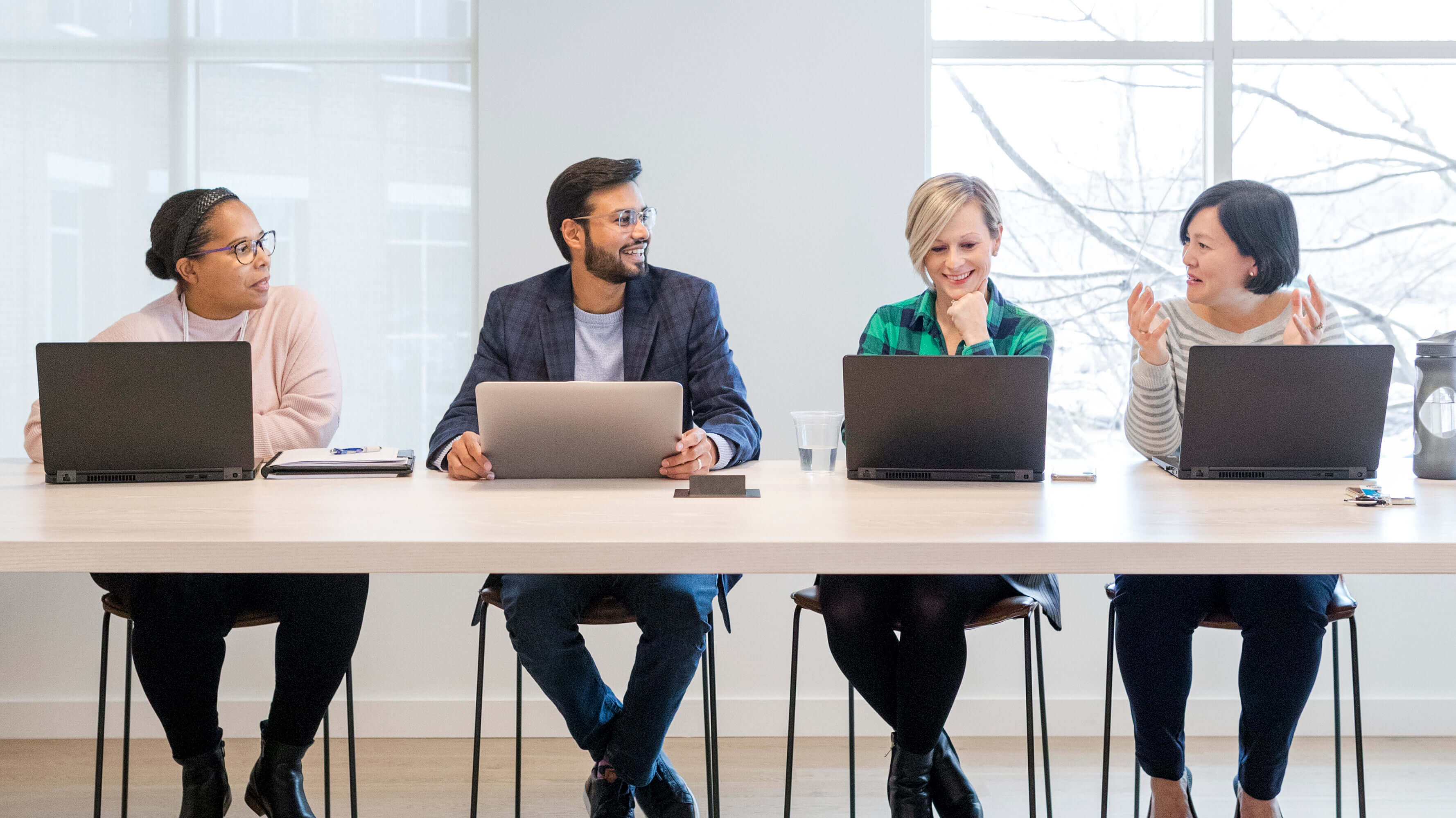 A long table, with a diverse group of people sitting there, have a lively and friendly discussion while using laptop computers.
