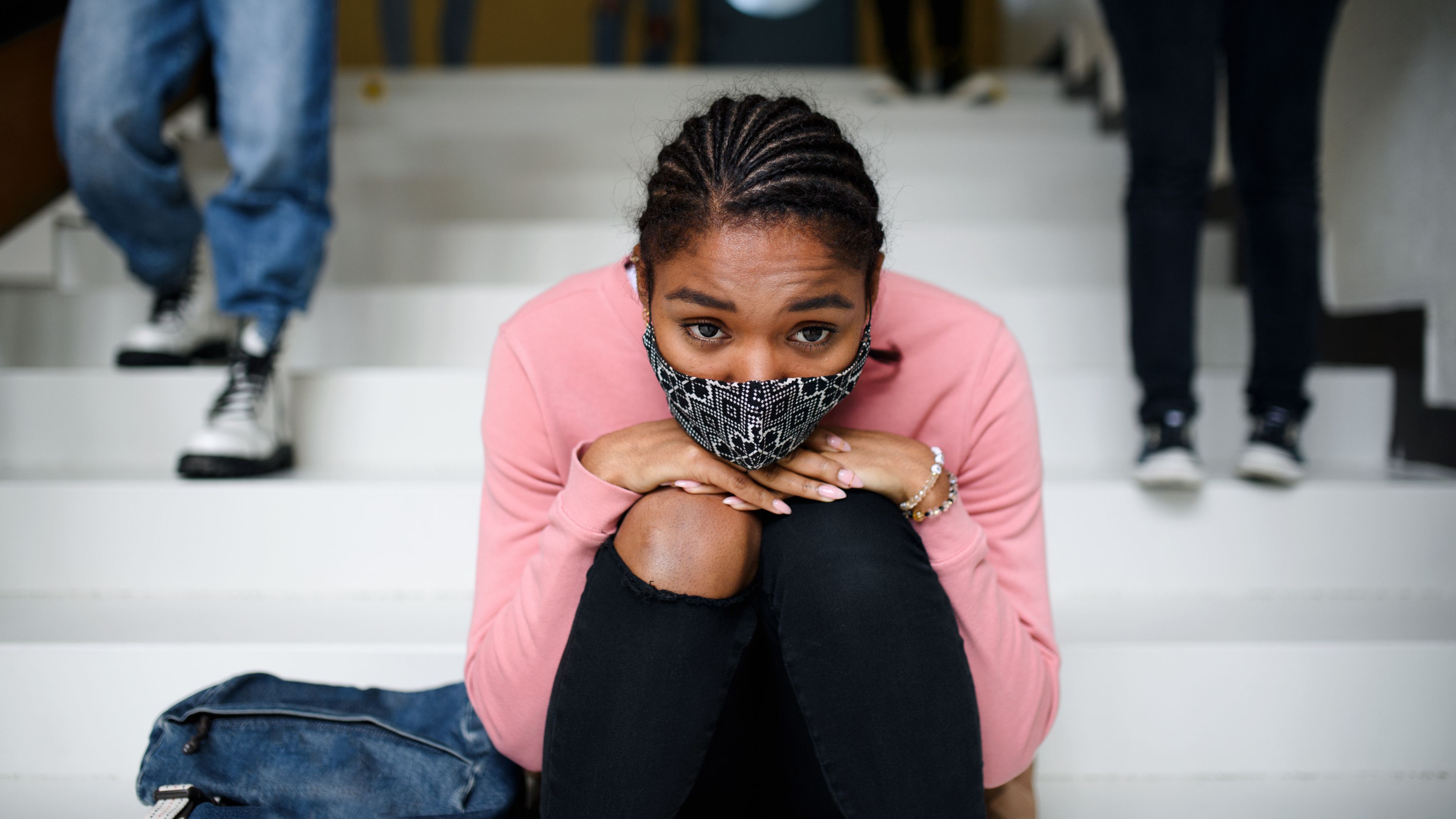A teenager wearing cornrows, a pink sweater and a mask sits on a staircase with her hands and chin on her knees while peers walk past.