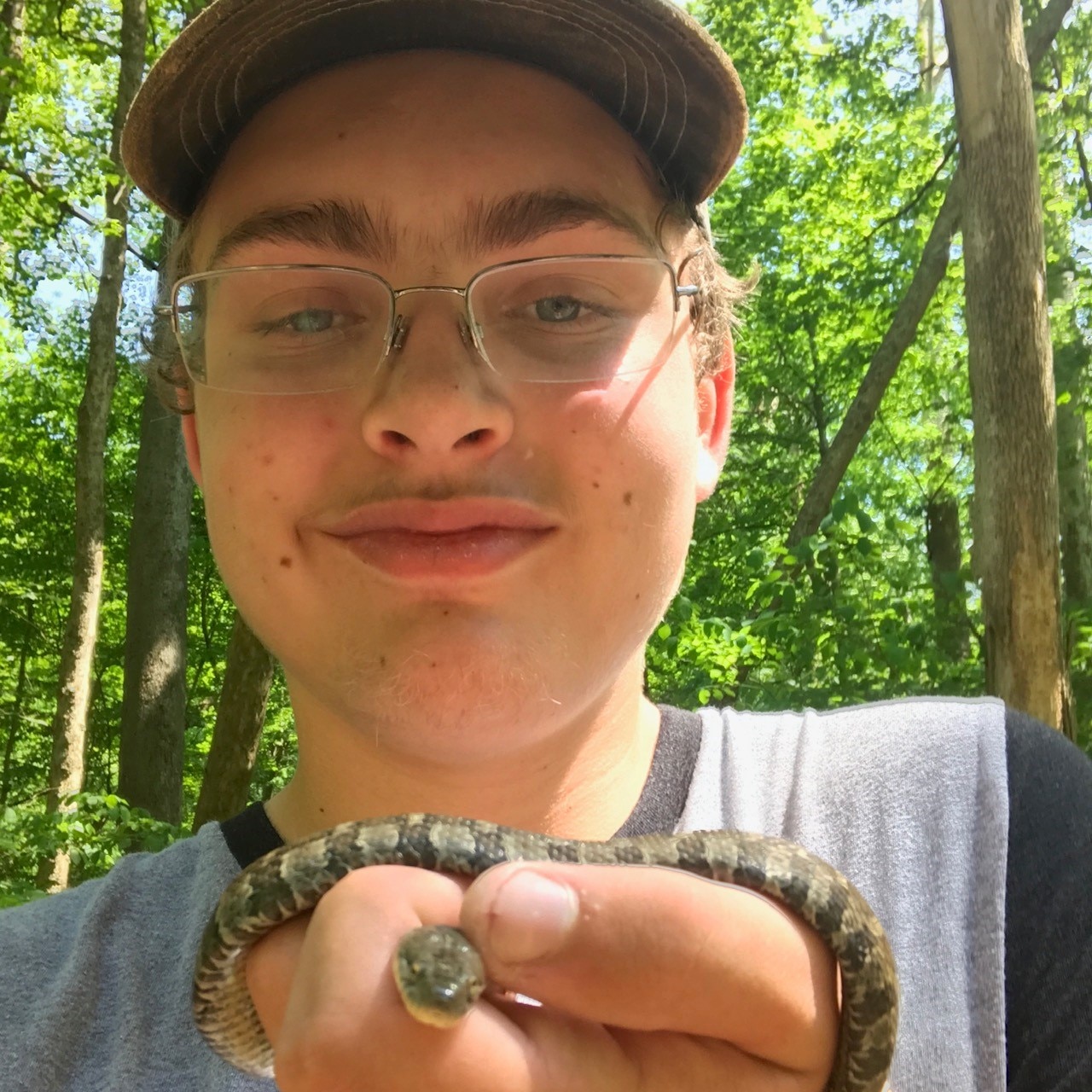 A teenage boy wearing a brimmed-hat, glasses, and a grey shirt smirks at the camera while holding a small snake in his hand.